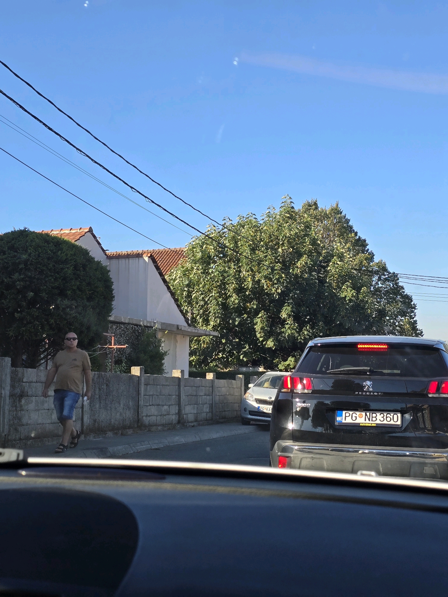 In Podgorica, Montenegro, a man wearing sunglasses, a tan t-shirt, blue shorts, and sandals walks on a sidewalk next to a gray brick wall. He is on the left side of the street, and behind him are trees and the roof of a building with red tiles. In front of the man, to his right, a black Peugeot SUV is stopped in traffic, its license plate reading "PG NB 360". To the left of the SUV is a silver car, partially visible, with a license plate beginning with "PG". The sky is clear and blue. The image appears to be taken from inside another vehicle, looking forward.
