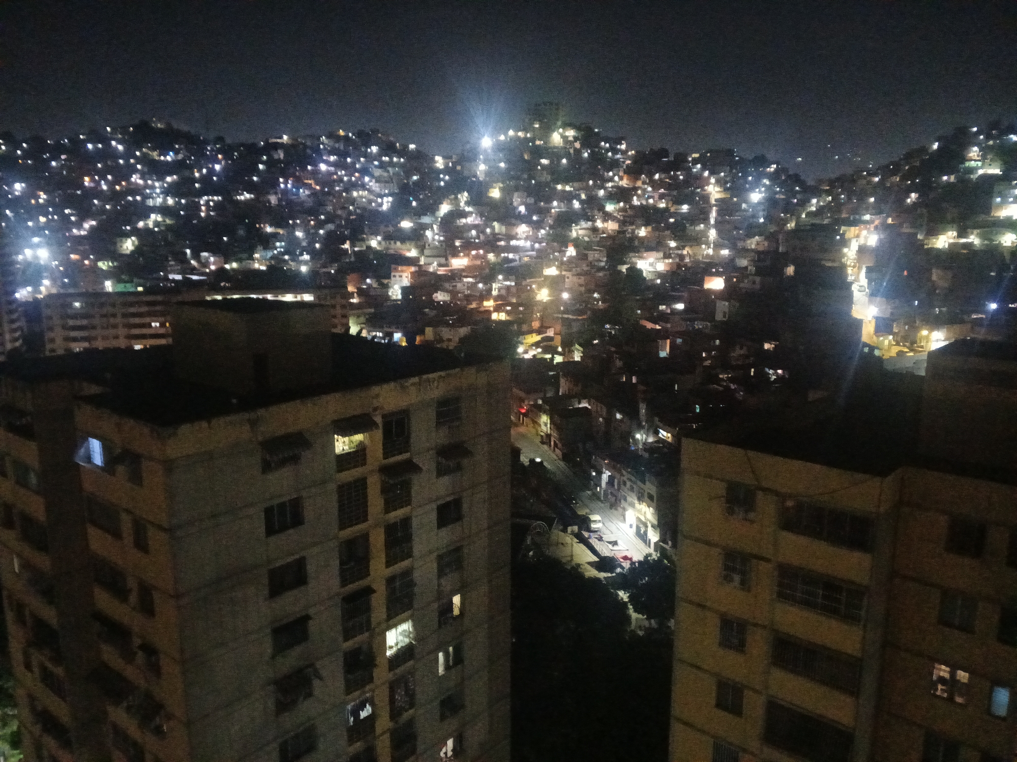 The image presents a nighttime view of Caracas, Venezuela, characterized by dense urban sprawl. In the foreground are two high-rise buildings, one grey and the other yellowish. Behind them, a hill is covered in densely packed housing illuminated by numerous lights, creating a visually striking pattern. These lights suggest a high population density in the hillside neighborhoods. A bright light can be seen near the top of the hill. The general impression is of an expansive urban landscape at night.