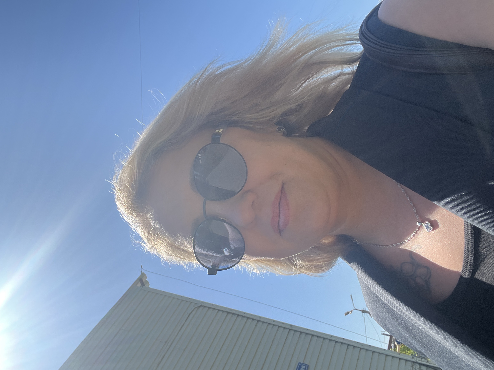 A woman with light blonde hair and sunglasses is taking a selfie outdoors on a sunny day. She is wearing a black top, and a silver necklace with a pendant. A tattoo is visible on her upper chest/neck area. In the background is a pale grey corrugated metal building. Above the building is a clear blue sky with a bright sun causing lens flare. The location is Chaikovsky, Russia, suggested by a cropped image and the context of the task.