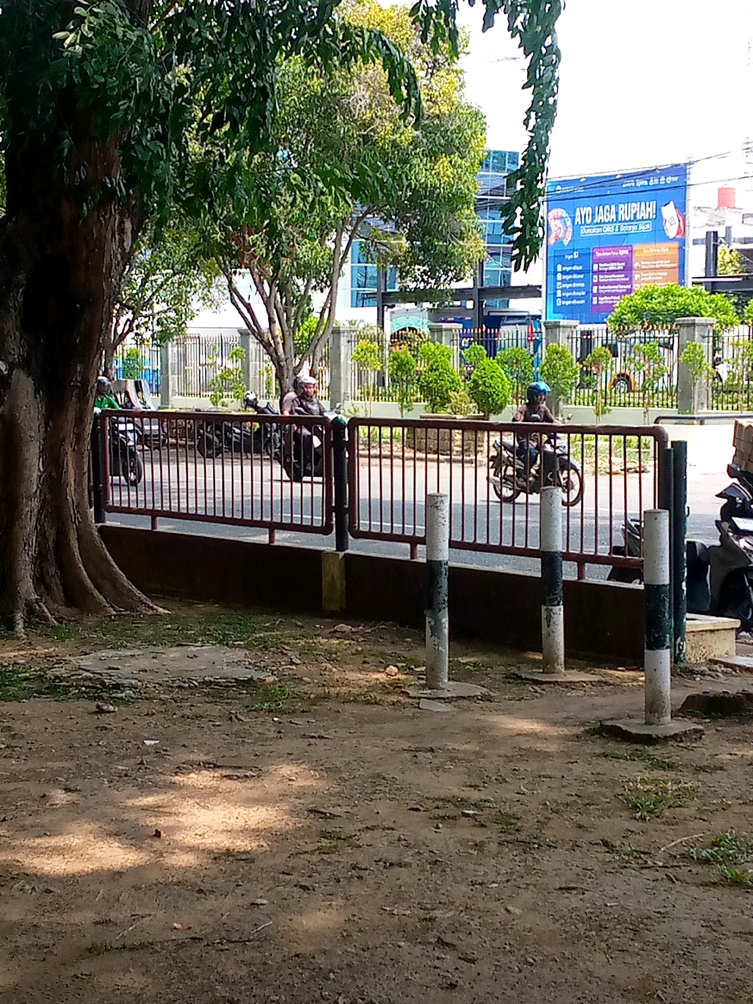 The image depicts a street scene in Lhokseumawe, Indonesia, likely near a commercial area. In the foreground, a large tree with rough bark is prominent on the left. The ground is a mixture of dirt, small stones, and sparse grass, with dappled sunlight indicating it's daytime. A red metal fence runs horizontally across the mid-ground, separating the viewer's area from the street.
Beyond the fence, several motorcycles are visible. Two individuals are riding motorcycles on the street. One rider, wearing a light-colored helmet and a dark shirt, is positioned in the right mid-ground, with their motorcycle partially obscured. Another rider, wearing a helmet and a dark shirt, is on a motorcycle in the left mid-ground. The street appears to be paved and has lane markings.
In the background, a modern building with a blue glass facade stands tall. A large blue banner with Indonesian text, including "AYO JAGA RUPIAH!", is prominently displayed on the building's exterior, suggesting a public awareness campaign. The banner has images and text related to currency and financial well-being. Other elements in the background include more fencing, decorative green shrubs, and the sky, which is bright and appears overcast or hazy. The overall impression is a busy urban environment with elements of nature and commerce.