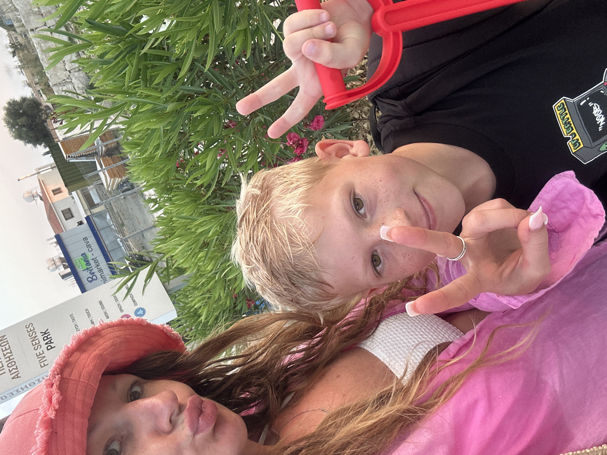 In Peyia, Cyprus, a woman wearing a pink bucket hat and pink clothing is taking a selfie with a boy. The boy, wearing a black t-shirt with a video game graphic, holds a red object and makes a peace sign. Behind them, a sign for "FIVE SENSES PARK" is visible, along with a "Britania minimarket - cava" store. Lush green foliage with small pink flowers surrounds them, and a residential building with solar panels can be seen in the background. The lighting suggests it is daytime.