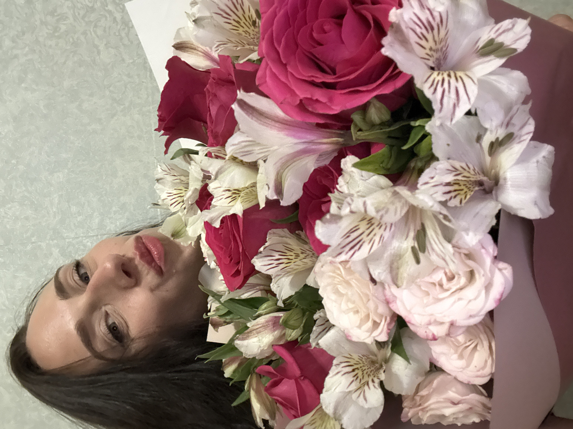 A woman lies on a light-colored surface, next to a bouquet of pink and white flowers, featuring roses and alstroemeria. Her face is close to the camera, showing dark hair, fair skin, and light pink lipstick. The background is a textured, light-colored surface. It's not possible to infer the location as the city of Saint Petersburg, Russia from the image.