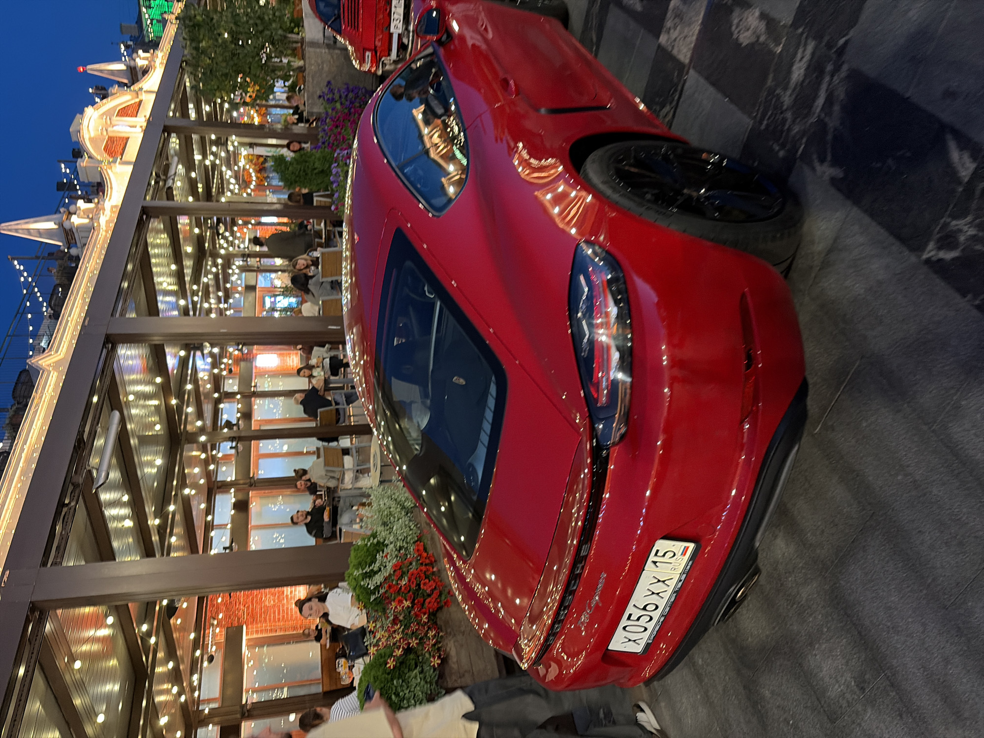 A bright red Porsche sports car is parked on a paved area in front of an outdoor seating area of a restaurant or cafe. The cafe has a covered roof supported by wooden beams and is illuminated by strings of lights. Several people are seated at tables in the cafe, some eating and some conversing. The background shows building facades, also adorned with lights, and a night sky. The license plate of the Porsche is visible and reads "X056XX 15 RUS". The scene appears to be a vibrant evening setting in Moscow, Russia.