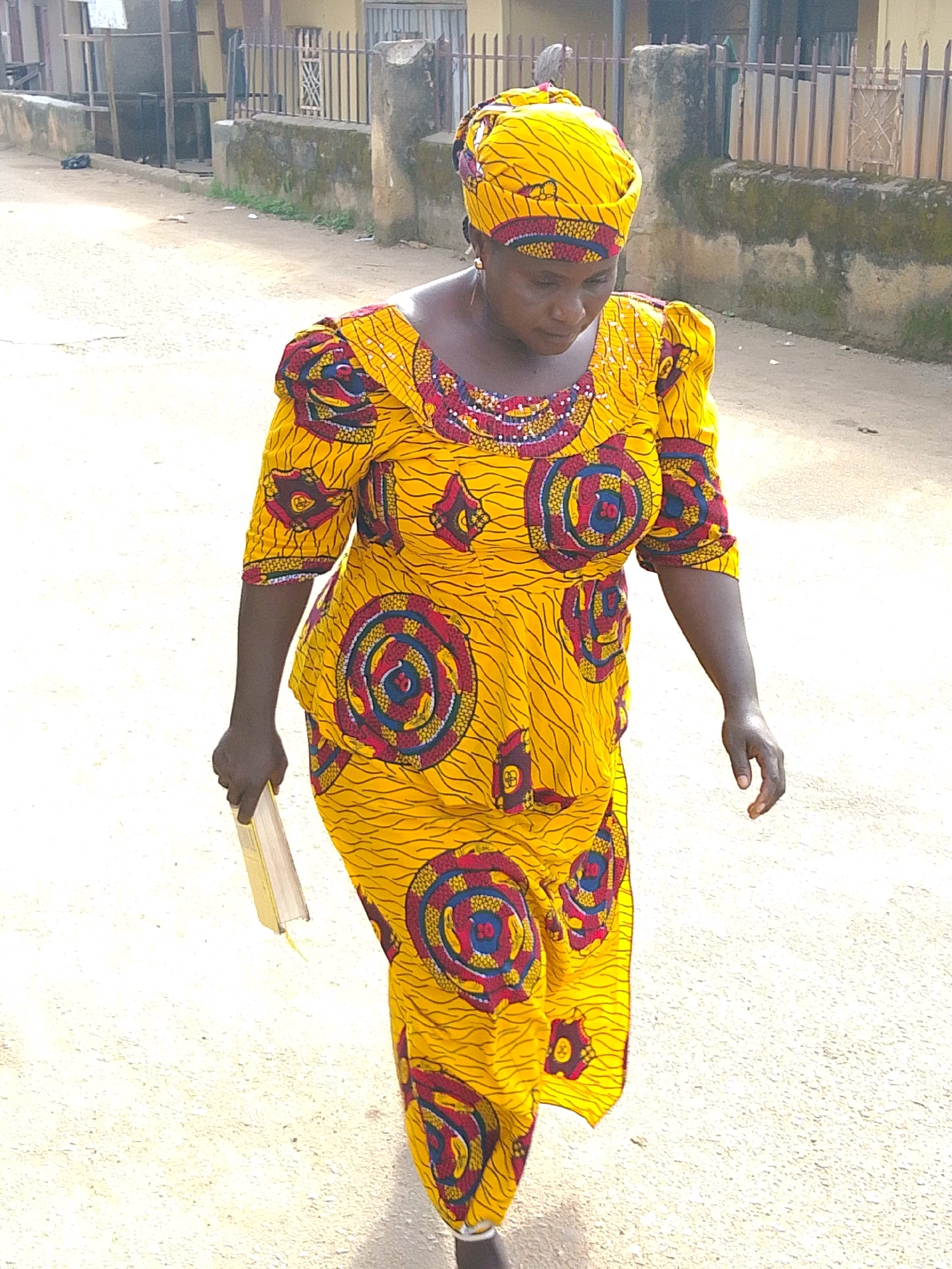 In the city of Jos, Nigeria, a woman wearing a bright yellow traditional African dress with a matching headscarf is walking on an unpaved road. Her dress is adorned with a vibrant red, blue, and yellow circular pattern and smaller red and blue geometric designs. The sleeves of her dress are puffed. She is holding a light-colored, rectangular object in her right hand. The background shows a street with buildings and a metal fence. The ground is dusty and appears to be a dirt road. The lighting suggests it is daytime, possibly late morning or early afternoon, with shadows cast on the ground.