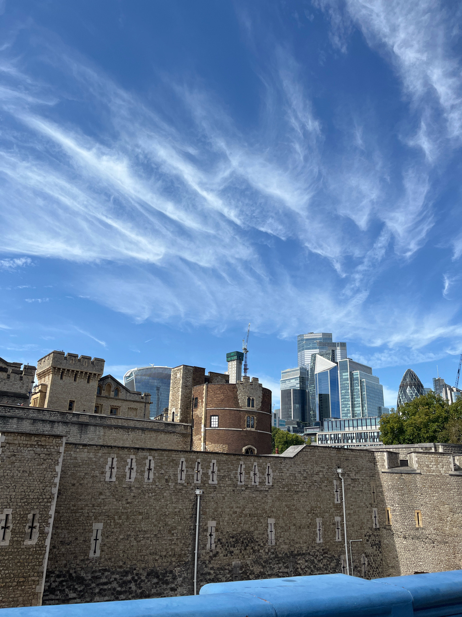 The image captures a view of the Tower of London with modern skyscrapers in the background, situated in the city of London, United Kingdom. The foreground is dominated by the stone walls and battlements of the Tower of London, characterized by their rough-hewn texture and crenellated tops. Small, narrow windows, likely for defensive purposes, are visible along the walls. A blue railing or barrier runs along the bottom of the frame, suggesting a viewpoint from a bridge or elevated walkway.
Behind the historic fortress, a collection of contemporary buildings rise into the sky. These include several glass-fronted skyscrapers with geometric designs, and the distinctive, rounded shape of the Gherkin building. The contrast between the ancient stone architecture of the Tower and the sleek, modern designs of the skyscrapers highlights the juxtaposition of old and new in London's urban landscape.
The sky above is a vibrant blue, streaked with wispy white clouds that form delicate, feathery patterns, indicating fair weather conditions. The lighting suggests it is daytime, with sunlight illuminating the scene and casting shadows. No people are explicitly visible in the frame, and the scene appears to be a static architectural and environmental study. The image composition emphasizes the historical depth of the Tower of London against the backdrop of its modern urban setting.