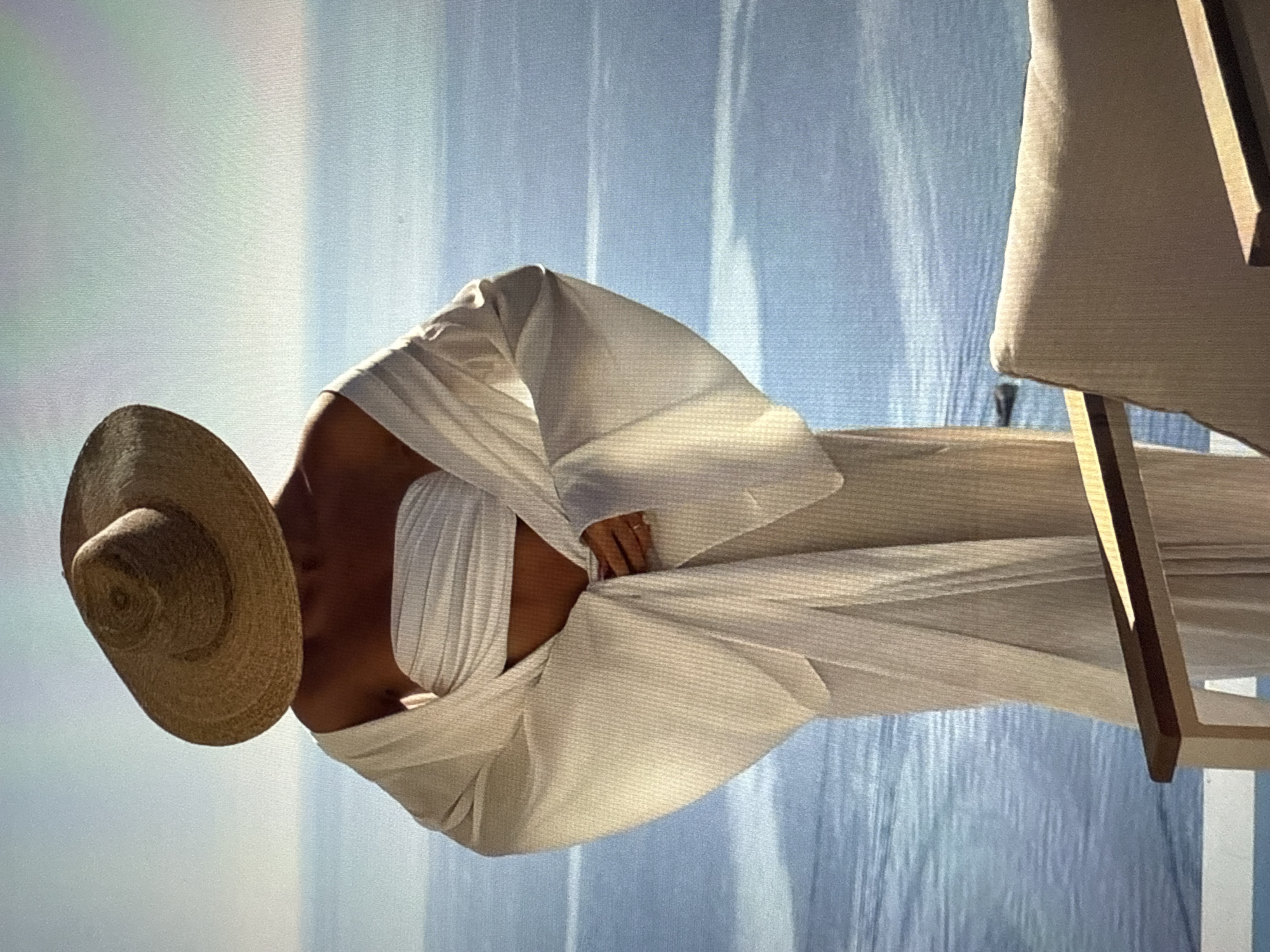 A person stands facing away from the camera, looking out at the ocean. They are wearing a large straw hat and a white bandeau top with a flowing white robe. The robe has wide, dramatic sleeves. The person's hands are clasped in front of them, and a ring is visible on one finger. The ocean is a deep blue, with some lighter patches indicating waves or reflections. The sky is a light blue with a hint of haze. In the foreground, the armrest of a chair is visible, made of wood and a light-colored fabric. The overall scene evokes a sense of tranquility and relaxation. This scene is not located in Găvănoasa, Moldova.
