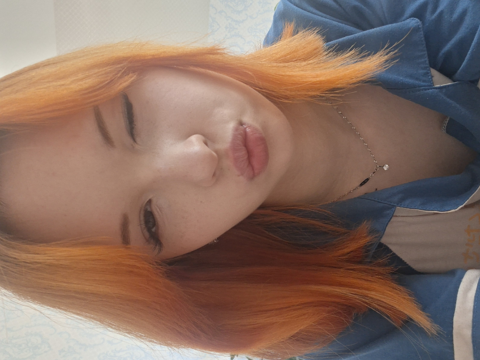 A close-up, tilted shot features a young woman with vibrant orange hair. Her face is predominantly in the frame, with one eye slightly closed, suggesting a wink or playful expression. Her lips are pursed as if posing for a kiss. She is wearing a blue top with white stripes on the sleeves and a delicate silver necklace with a small pendant. Her skin is fair and has a few visible freckles. The background is a softly patterned wallpaper, suggesting an interior setting. There is no indication of the location being Chaikovsky, Russia.
