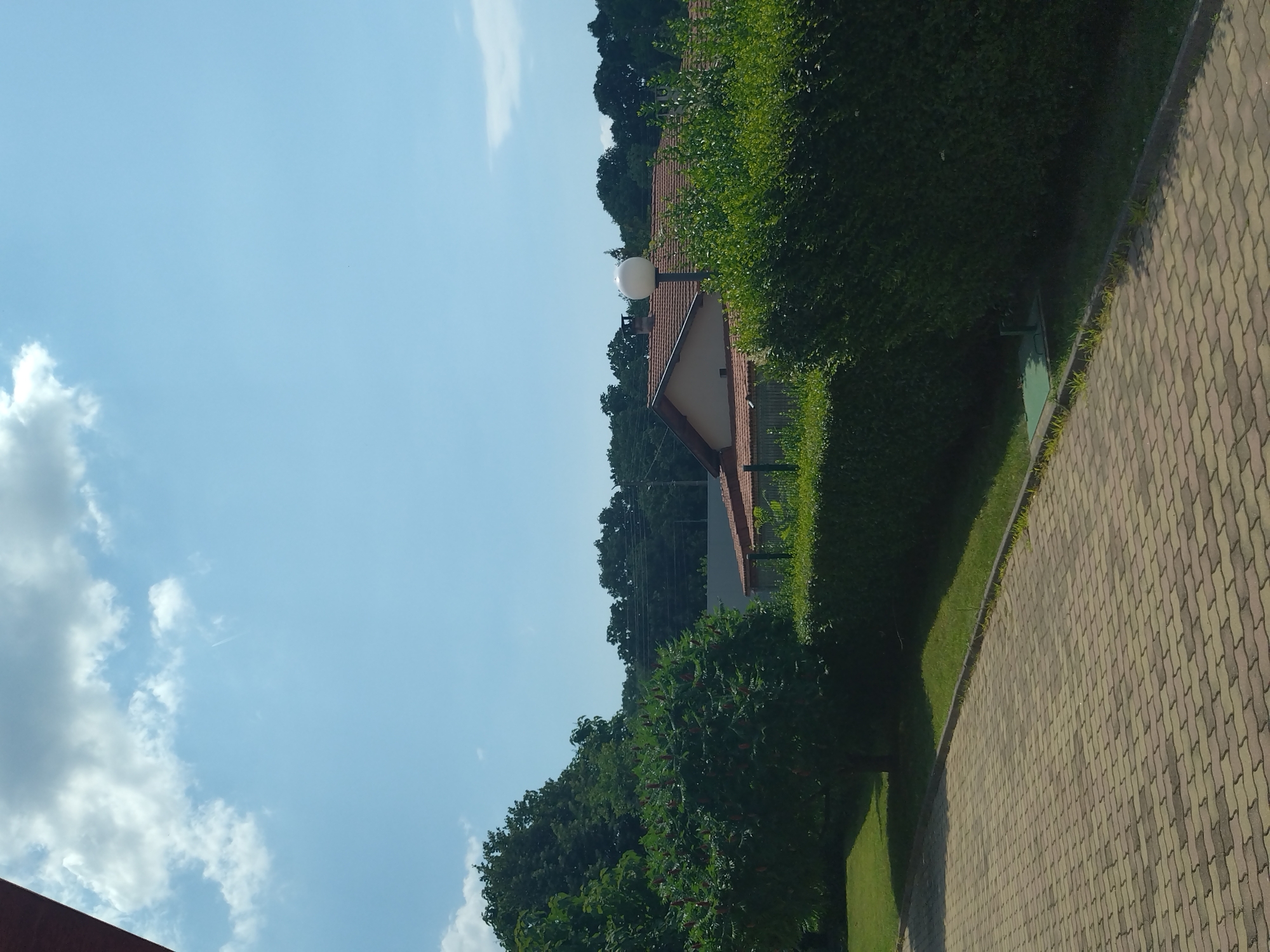 The image depicts a suburban scene in Yagoda, Bulgaria. In the foreground, a paved walkway composed of interlocking gray and beige bricks leads towards the viewer. To the right of the walkway, a meticulously trimmed green hedge forms a boundary. Beyond the hedge, the roof of a building with terracotta tiles is visible, partially obscured by more greenery. A spherical white outdoor light fixture stands on a pole in front of the building. In the middle ground, dense green trees and foliage create a lush backdrop. A tall, dark green tree with reddish-brown clusters is prominent on the left. The background features a line of trees against a clear blue sky with scattered white clouds. The scene suggests a well-maintained residential area on a sunny day.