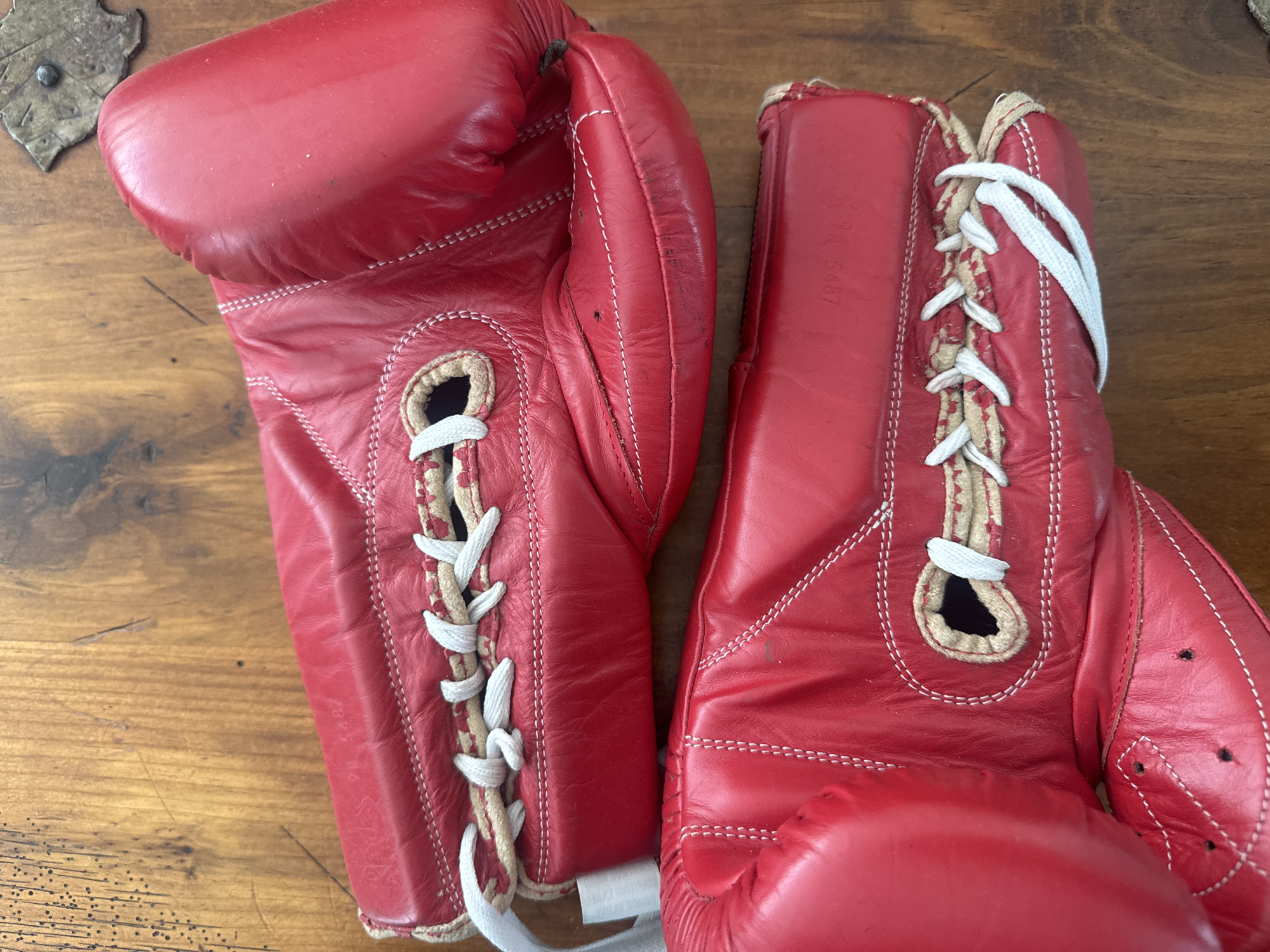 The image shows a pair of vintage red leather boxing gloves lying on a wooden surface. The gloves are laced with white cord. The right glove is visibly worn, with a faint inscription that appears to read "71 5487". No people, actions, or interactions are depicted. The environment is limited to the wooden surface and the gloves themselves. There is no information in the image to indicate a specific location in Puebla, Mexico, or any city.