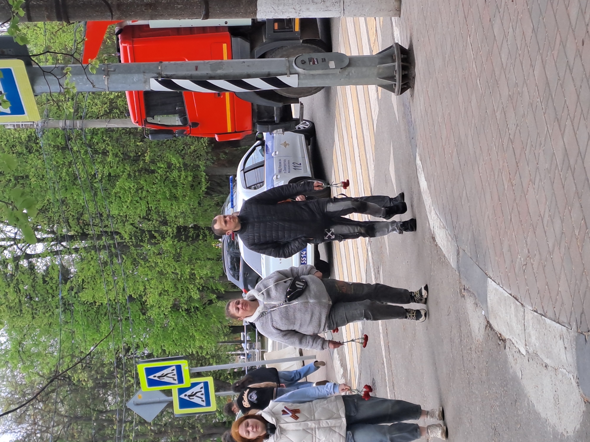 The image shows a street scene in Elektrostal, Russia. Several people are walking across a pedestrian crosswalk, each holding a small bunch of red flowers. A police car is parked on the street behind the crosswalk, and a large red truck is partially visible in the background. A street sign with a pedestrian crossing symbol is visible on the left side of the frame. The sidewalk is paved with brick. Trees with green foliage are visible in the background.
