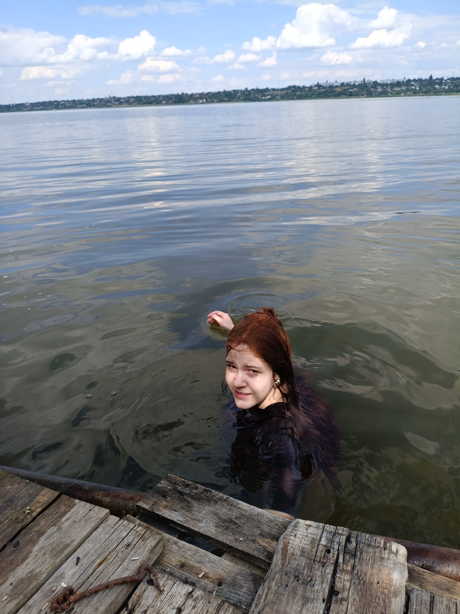 A young woman with auburn hair is swimming in a body of water, likely a river or lake. She is partially submerged, wearing a dark-colored garment that appears to be wet. Her face is visible, and she is looking towards the camera with a slight smile. In the foreground, there is a wooden structure, possibly a dock or pier, made of weathered planks. The water has ripples and reflections of the sky. In the distance, a treeline and buildings suggest a settlement. The sky above is blue with scattered white clouds. The overall scene depicts a natural outdoor setting, likely during daylight hours.