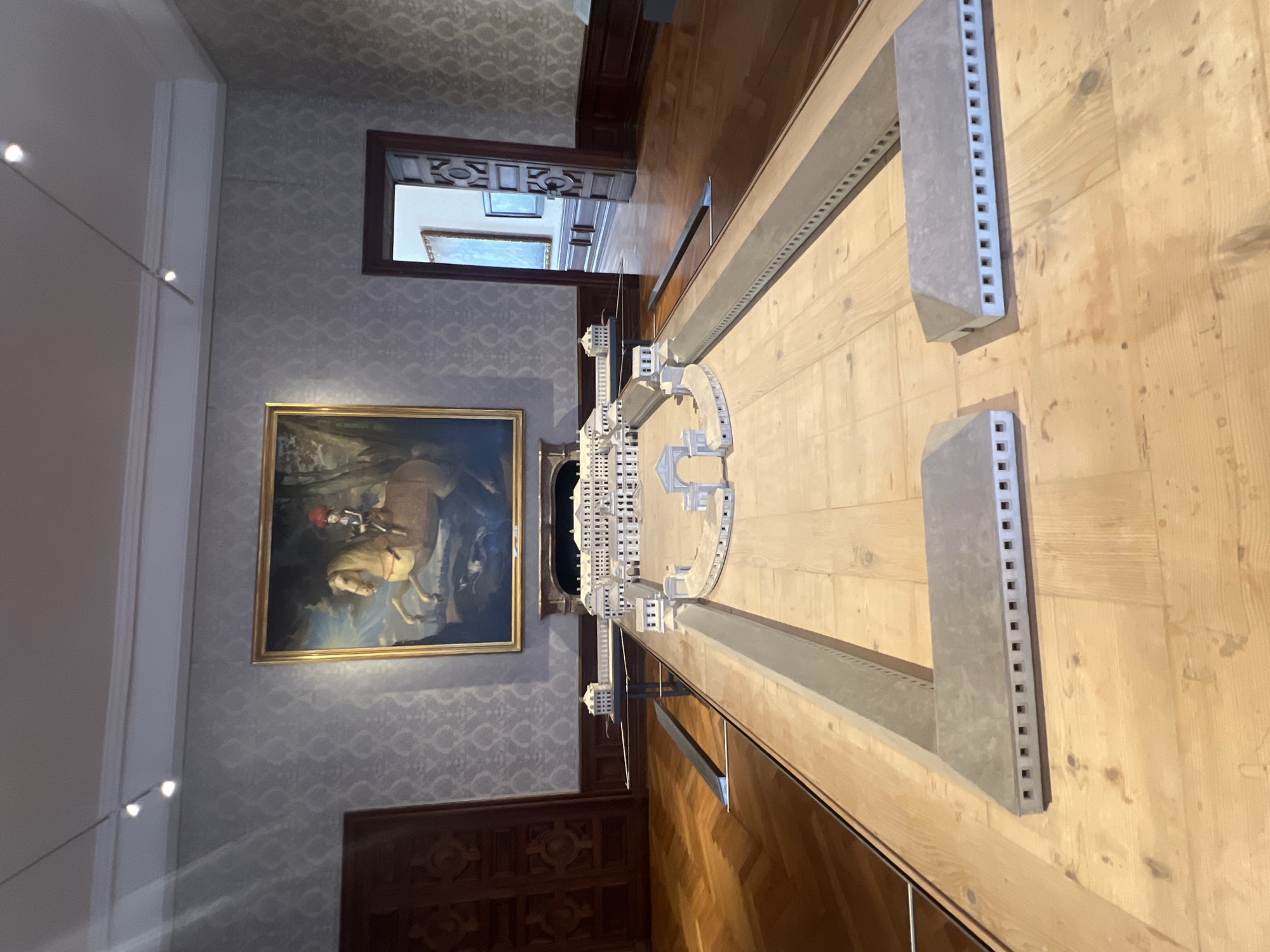 This image shows a model of the Schleissheim Palace complex in Oberschleißheim, Germany. The model is displayed on a wooden table in a museum setting. The model depicts the main palace buildings, courtyards, and surrounding structures in a detailed, architectural style. In the background, a large painting of a man on horseback hangs on a wall. The room has wood-paneled walls and a parquet floor. The overall impression is of a well-preserved historical site being showcased.
