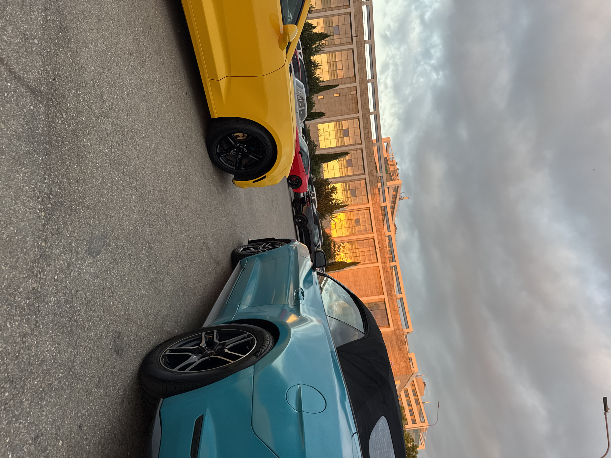The image captures a parking lot scene with several cars, including a yellow Chevrolet Camaro in the foreground on the left and a teal convertible Ford Mustang in the foreground on the right. In the background, a long, modern building with large glass windows reflects the warm, golden light of the setting sun. The sky is filled with dramatic clouds. The overall environment suggests an outdoor event or gathering of car enthusiasts, possibly in the city of Sirius, Russia, given the architectural style and lighting conditions often associated with that region during sunset.