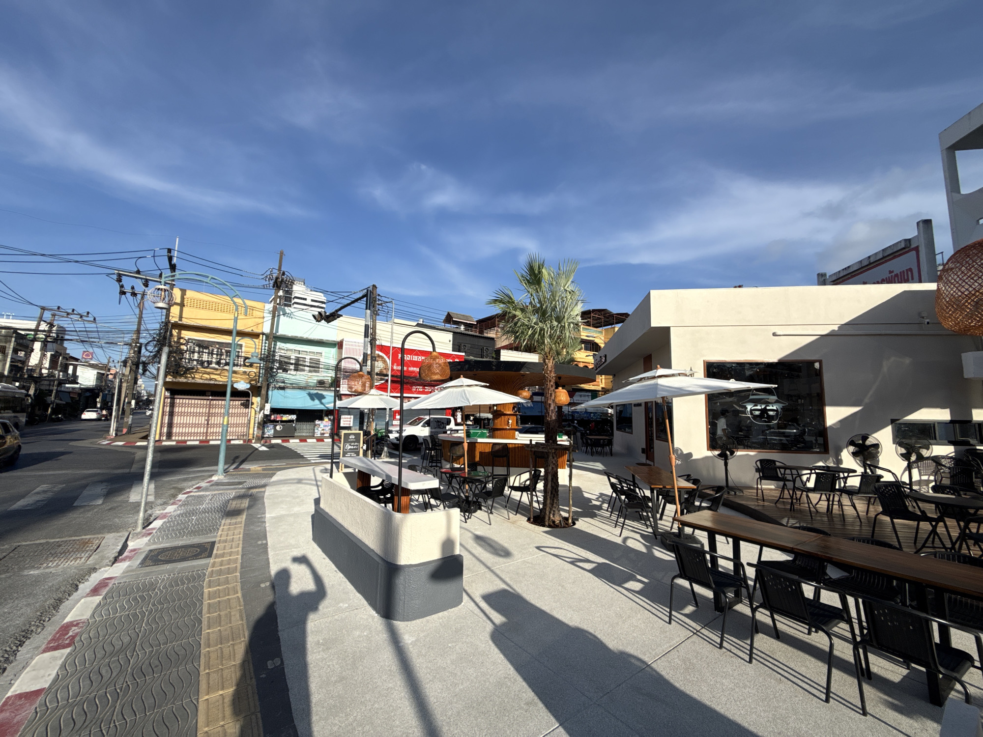 This is a wide-angle, eye-level, outdoor, daytime photograph of a street scene in Phuket City Municipality, Thailand. The foreground features a paved sidewalk with tactile paving and a streetlamp. A person's shadow is visible on the sidewalk. The middle ground shows an outdoor seating area for a restaurant or cafe, with tables, chairs, and umbrellas. A palm tree stands in the center of the seating area. In the background, there are buildings, a street with a crosswalk, and electrical wires. The sky is blue with some clouds. The buildings in the background are a mix of styles and colors, with some appearing to be older shophouses. There is a yellow building with blue trim and a light green building with a red sign. A white building with large windows is prominent on the right side of the image. A few vehicles are visible on the street. The scene depicts a bustling urban environment with commercial activity.