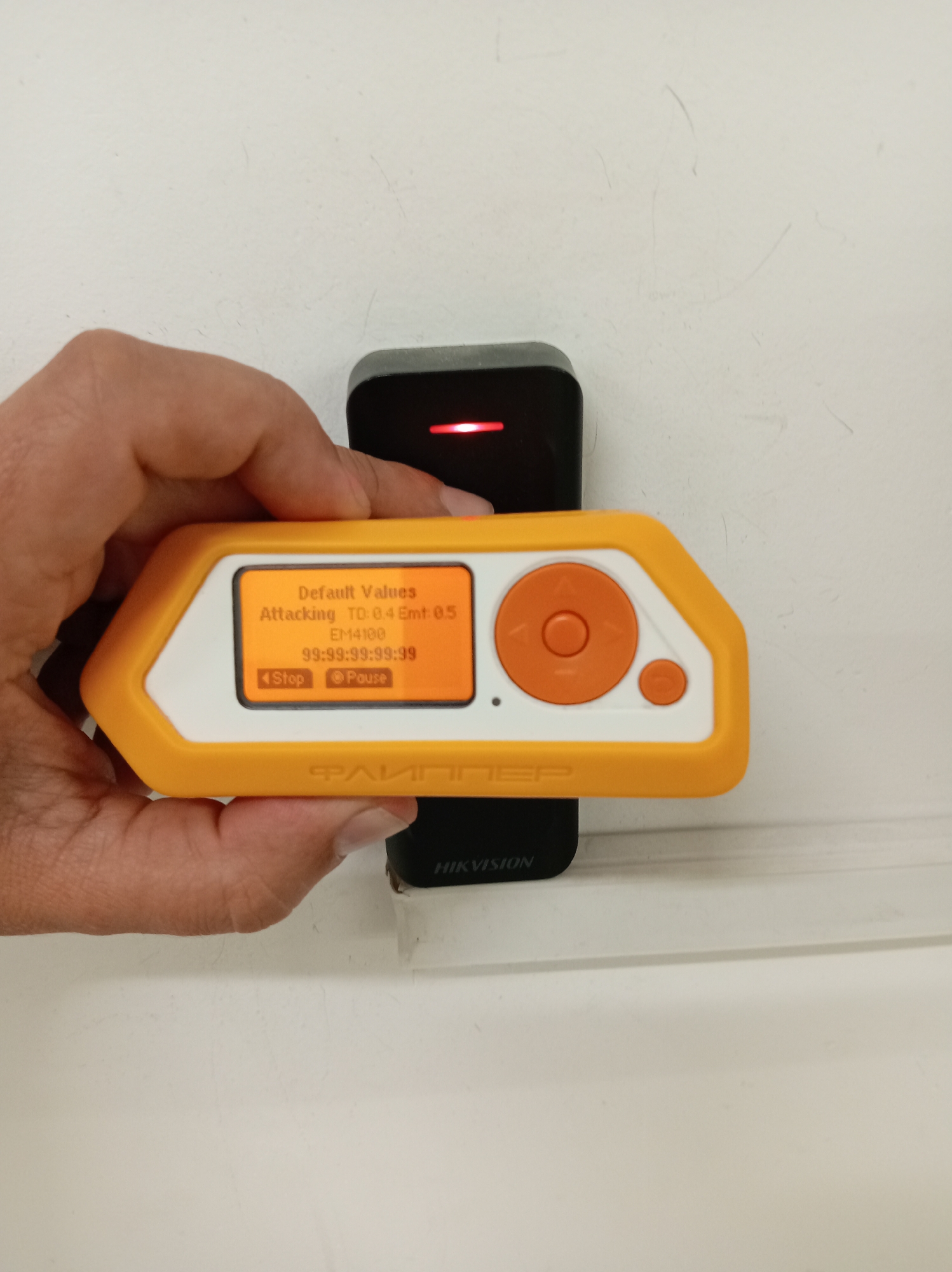 A hand holds an orange and white electronic device with a screen displaying "Default Values", "Attacking TD: 0.4 Emt: 0.5", "EM14100", and "99:99:99:99:99". Below this text are two buttons labeled "Stop" and "Pause". The device is positioned in front of a black electronic component with a red light. The background is a white, textured wall. The bottom of the black component shows the "HIKVISION" logo. The scene appears to be indoors.