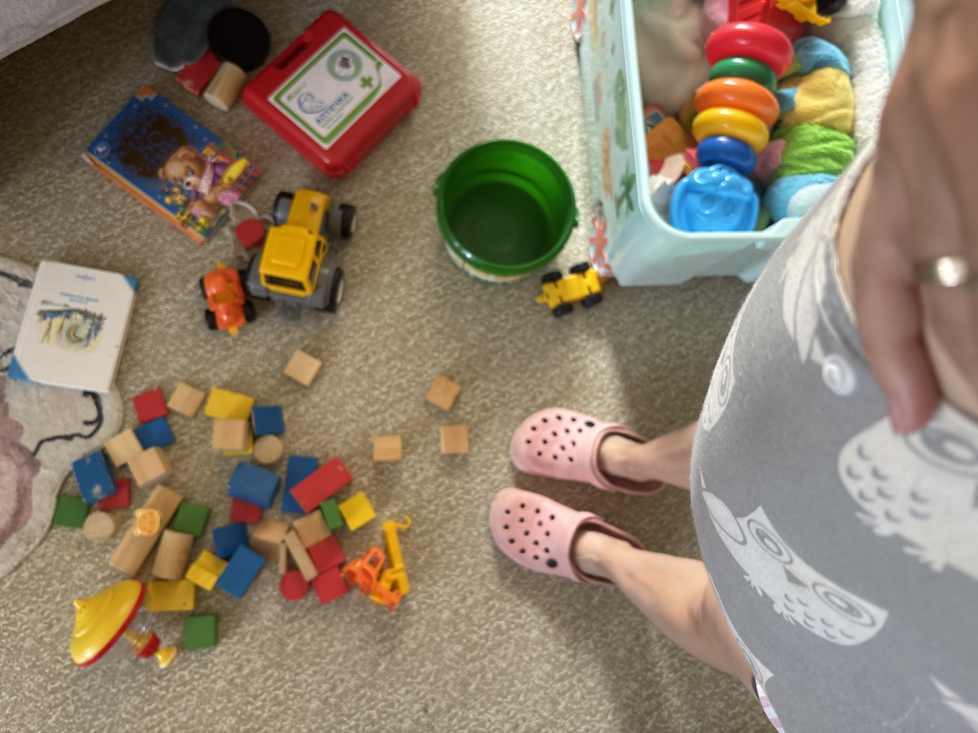 The image shows a top-down view of a person's feet wearing pink Crocs, positioned on a light-colored carpet. Scattered around the feet are various children's toys, including wooden blocks of different colors, a yellow spinning top, a yellow and grey toy truck, a red toy car, a green bucket, and a blue and white toy chest filled with colorful stacking rings and other toys. In the background, there are two children's books, one with a teddy bear illustration. The scene appears to be a play area within a residential setting. There is no discernible information in the image that would allow for the determination of its location within Naberezhnye Chelny, Russia.