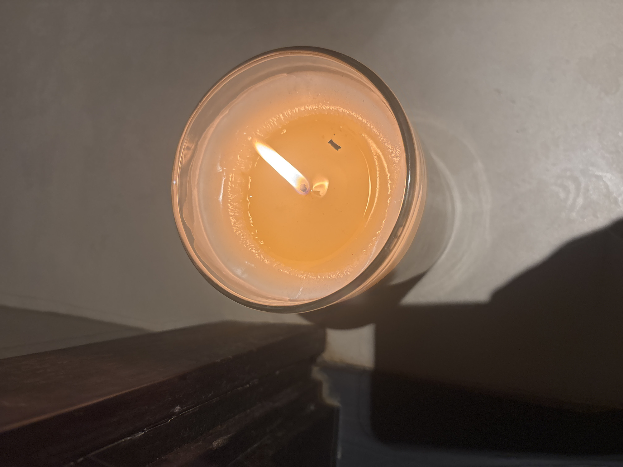 The image shows a top-down view of a lit candle in a glass jar. The candle wax is melted and a vibrant orange color, with a bright yellow flame. A small black speck, possibly a piece of the wick, is visible on the surface of the wax. To the left of the candle, a dark wooden surface casts a shadow on the light-colored ground. The lighting suggests an indoor environment, likely in the evening or a dimly lit room. There is no indication of the city of Dubai, United Arab Emirates, or any specific location within it. The scene is devoid of people or any interactions.