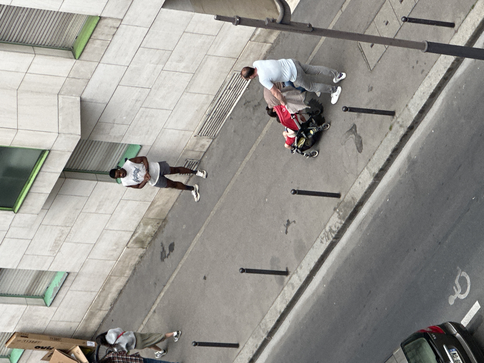 Aerial view of a sidewalk and roadway in Paris, France. On the sidewalk, three men are visible: One man is wearing a white t-shirt and grey pants, tending to a stroller. Another man wears a white tank top and grey shorts. A third man wears a grey long sleeve shirt, plaid shorts, and is sitting on the ground near some boxes. A black vehicle is parked on the road, next to a handicapped parking symbol. Black bollards line the sidewalk. A building is adjacent to the sidewalk, featuring window grates and green window frames.