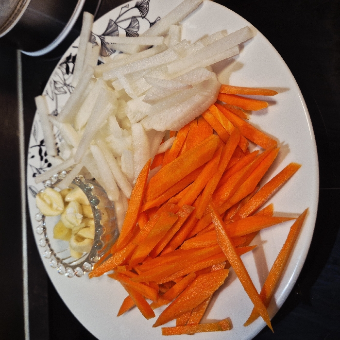 The image displays a white, oval-shaped plate filled with pre-cut vegetables, suggesting ingredients prepared for cooking. The plate itself has a decorative black and white pattern resembling Ginkgo leaves on its upper left side.
On the plate, two main types of julienned vegetables are neatly separated. On the left and upper-middle section, there is a pile of white, thinly cut strips, characteristic of daikon radish or similar root vegetable. To the right and lower-middle, a vibrant orange pile consists of thinly julienned carrots. Both vegetables are uniformly cut into long, slender sticks.
In the lower-left quadrant of the plate, nestled between the white vegetable strips and the edge of the plate, is a small, clear, decorative glass bowl. This bowl contains several peeled garlic cloves, some whole and some halved. The rim of the glass bowl has a textured or beaded design.
The background of the image is dark, likely a kitchen countertop or stovetop, with a hint of a metallic pot or pan visible in the upper-left corner, out of focus. The lighting appears to be indoor and consistent, illuminating the vegetables clearly. The overall impression is one of meticulous preparation of ingredients for a meal.