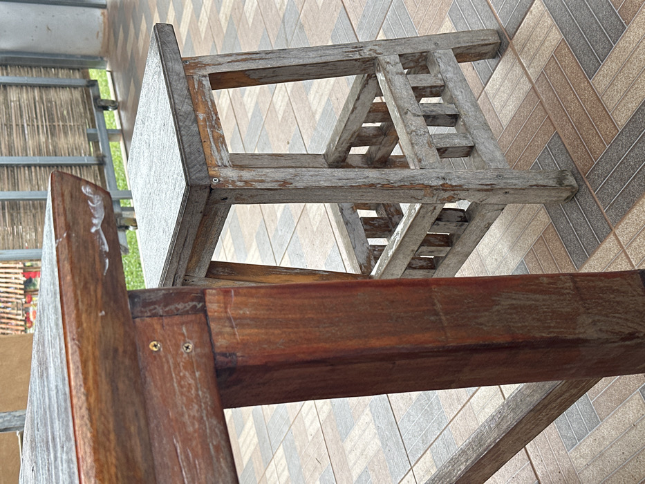 The scene is set in a semi-outdoor patio area in Ban Mai Saha Sampan, Thailand, under daylight conditions. The foreground features two weathered wooden furniture pieces. On the left, a reddish-brown wooden table with a rectangular top and robust square legs displays visible screws securing its structure. The table's top edge exhibits several white, irregular marks. To the right, a gray-toned wooden stool with a square seat and four legs stands adjacent. This stool includes a lower rectangular frame constructed from horizontal and vertical wooden slats, serving as a footrest or brace, with visible screws in its construction. Both the table and stool show signs of significant weathering, with faded finishes and surface wear. The floor beneath the furniture consists of multi-toned rectangular ceramic tiles arranged in a staggered or herringbone pattern, incorporating shades of brown, gray, and beige. The background, out of focus, reveals a metal railing or frame supporting horizontal bamboo or wooden screen elements, with green foliage visible beyond. No individuals are present in the image, and no active events are occurring.