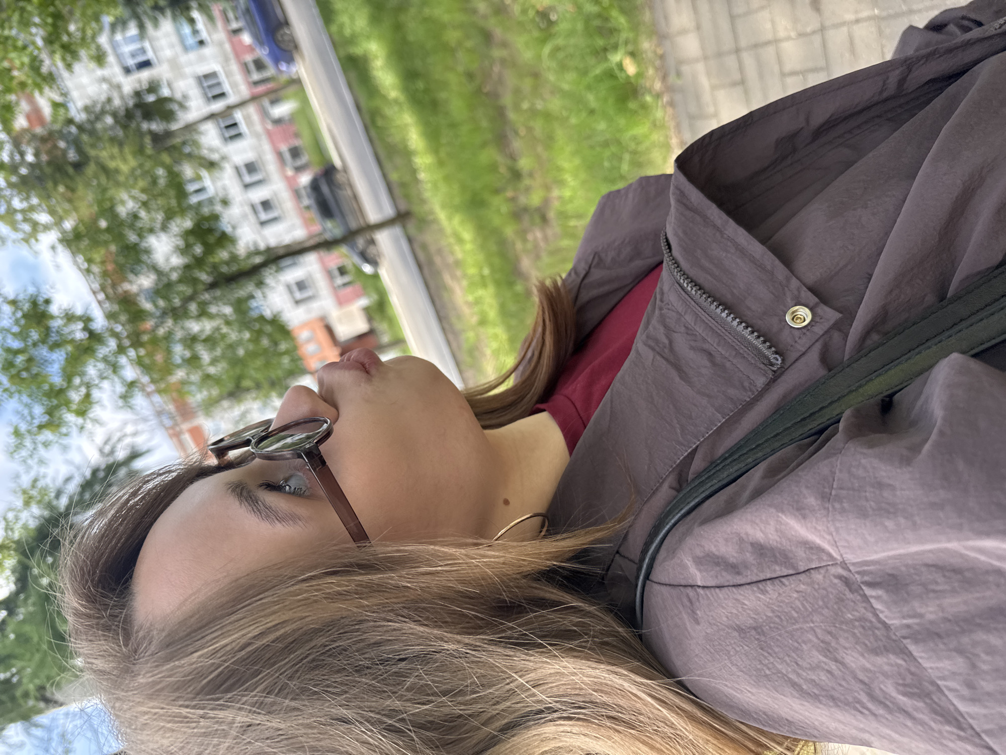 A young woman with blonde hair and glasses is captured in a close-up, side-profile shot. She is wearing a dark purple jacket over a maroon shirt and has a dark bag slung across her shoulder. The background suggests an urban setting with a building, trees, and a road with cars. The image appears to be taken outdoors in Chaikovsky, Russia, given the presence of typical Russian apartment buildings and the style of cars visible. The woman is looking upwards and to the left with her lips pursed.
