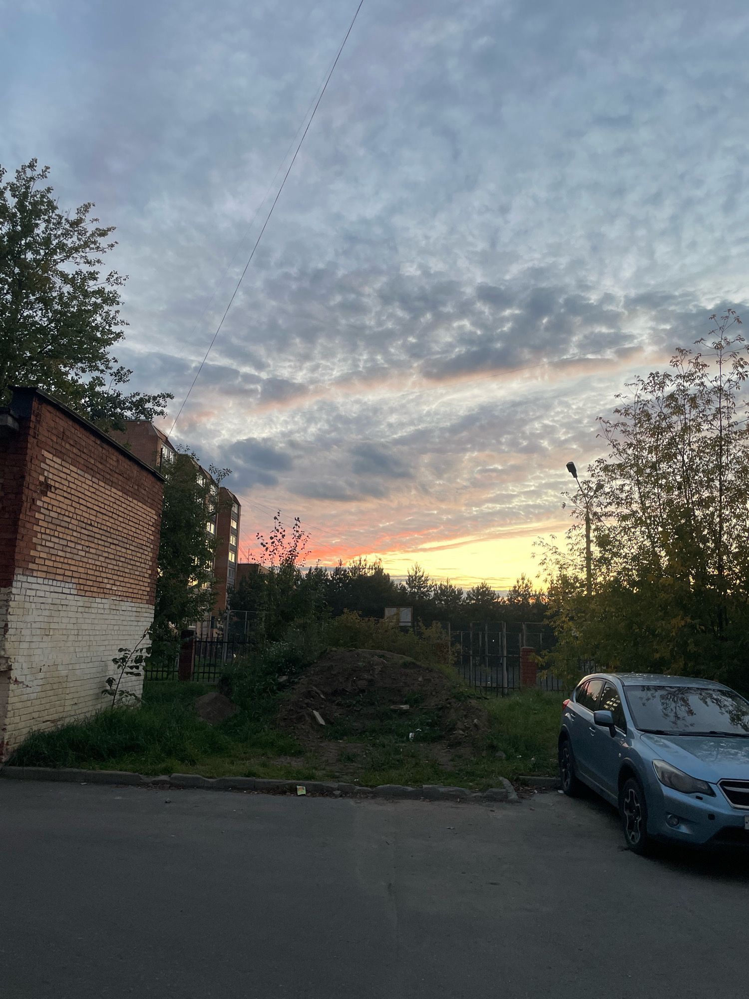 The image depicts an exterior scene in the city of Selyatino, Russia, likely at dusk or dawn. The sky is filled with dramatic, textured clouds, with hints of pink and yellow suggesting the presence of a sunset or sunrise. In the foreground, a paved road occupies the lower portion of the frame. To the left, a weathered brick building with white trim is visible, partially obscured by trees. Directly behind the building, a multi-story apartment complex stands, its windows reflecting the sky. A pale blue SUV is parked on the right side of the road, facing away from the camera. Dense vegetation, including trees and tall grass, lines the background, with a dark metal fence visible behind the car and extending into the distance. A lamppost stands amongst the trees to the right of the apartment building. A pile of dark soil is situated on the grassy area between the road and the fence. No people are present in the image.