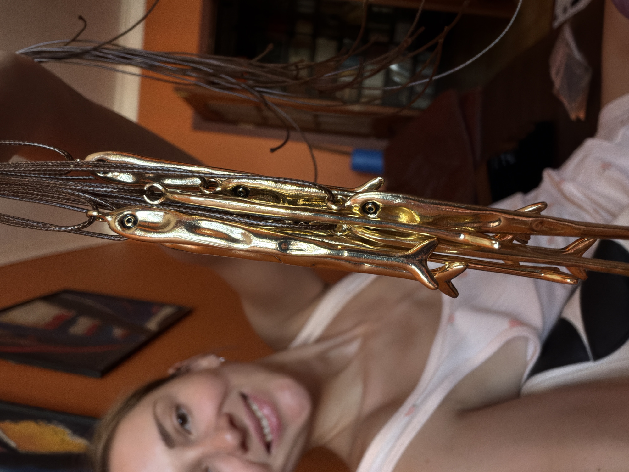 A close-up, waist-up shot shows a woman holding a cluster of golden pendants on thin brown cords. The pendants are elongated, stylized human figures with bent limbs. The woman, with blonde hair, is smiling broadly and wearing a white tank top with a subtle pink pattern. Her left ear is visible, adorned with a small stud earring. The background consists of an orange wall, two framed pictures, and a dark wooden surface with some indistinct objects. A bright blue cylindrical object is visible on the wooden surface. The scene appears to be indoors, possibly a room in a home or a shop. The image is not taken in Khimki, Russia.