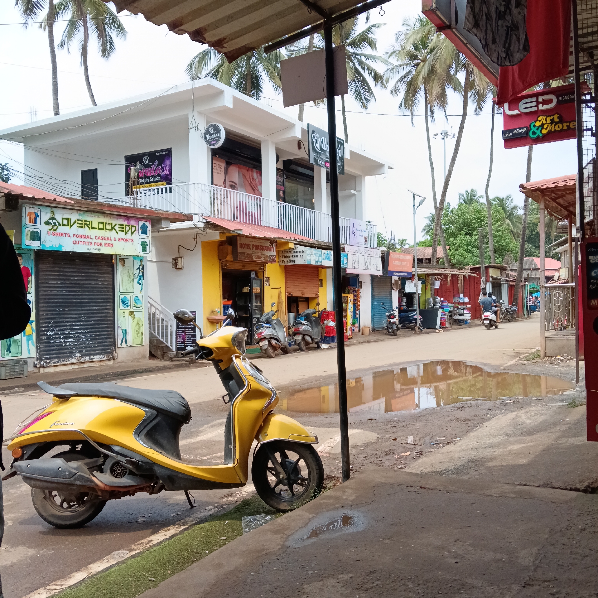 The image depicts a street scene in Oxel, India. In the foreground, a yellow scooter is parked on a concrete surface near a patch of grass and a puddle. A person partially visible on the left is out of focus.
Behind the scooter, the street leads past a row of small businesses. On the left, a shop named "Overlockedd" with closed shutters is visible. Adjacent to it is a two-story white building with balconies and signage for "Urmila's Beauty Saloon". Next to that is "Hotel Prabhavati".
Across the street, a sign reads "LED Art & More". Further down the street, people on scooters and pedestrians can be seen moving along the road. Palm trees are visible in the background, giving the scene a tropical feel. The road is partly wet with puddles of water.