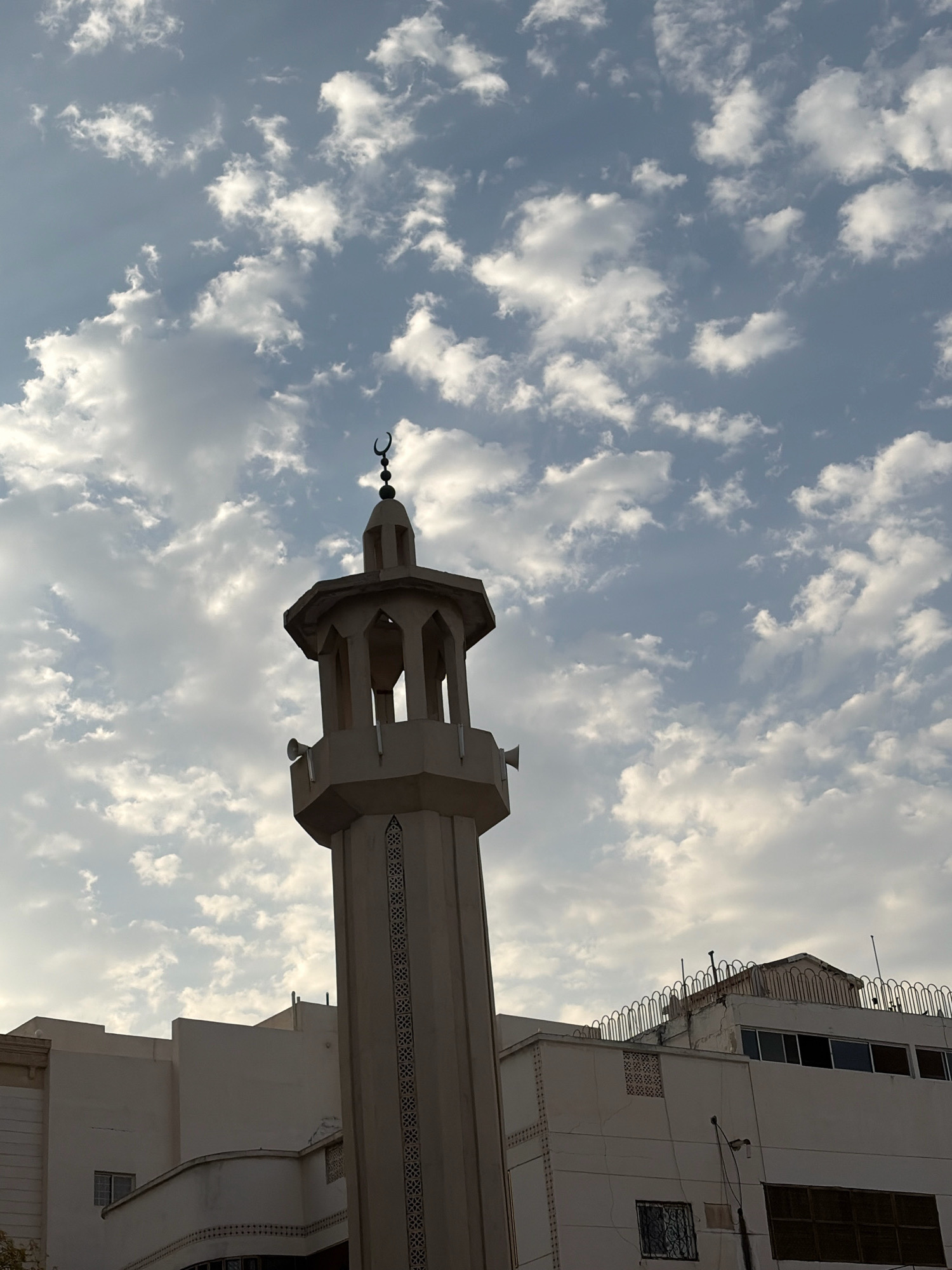 The image shows a minaret of a mosque in Medina, Saudi Arabia, against a sky filled with white clouds. The minaret is tall and slender, with a distinctive Islamic architectural style. It has a hexagonal base and is topped with a smaller, open structure that houses speakers. A crescent moon is visible at the very top of the minaret. In the background, several modern buildings are visible, suggesting an urban setting. The buildings are predominantly white and appear to be several stories high. The sky is a light blue, with scattered clouds that create a dramatic and textured backdrop. The lighting suggests it is either early morning or late afternoon. There are no people visible in the image.