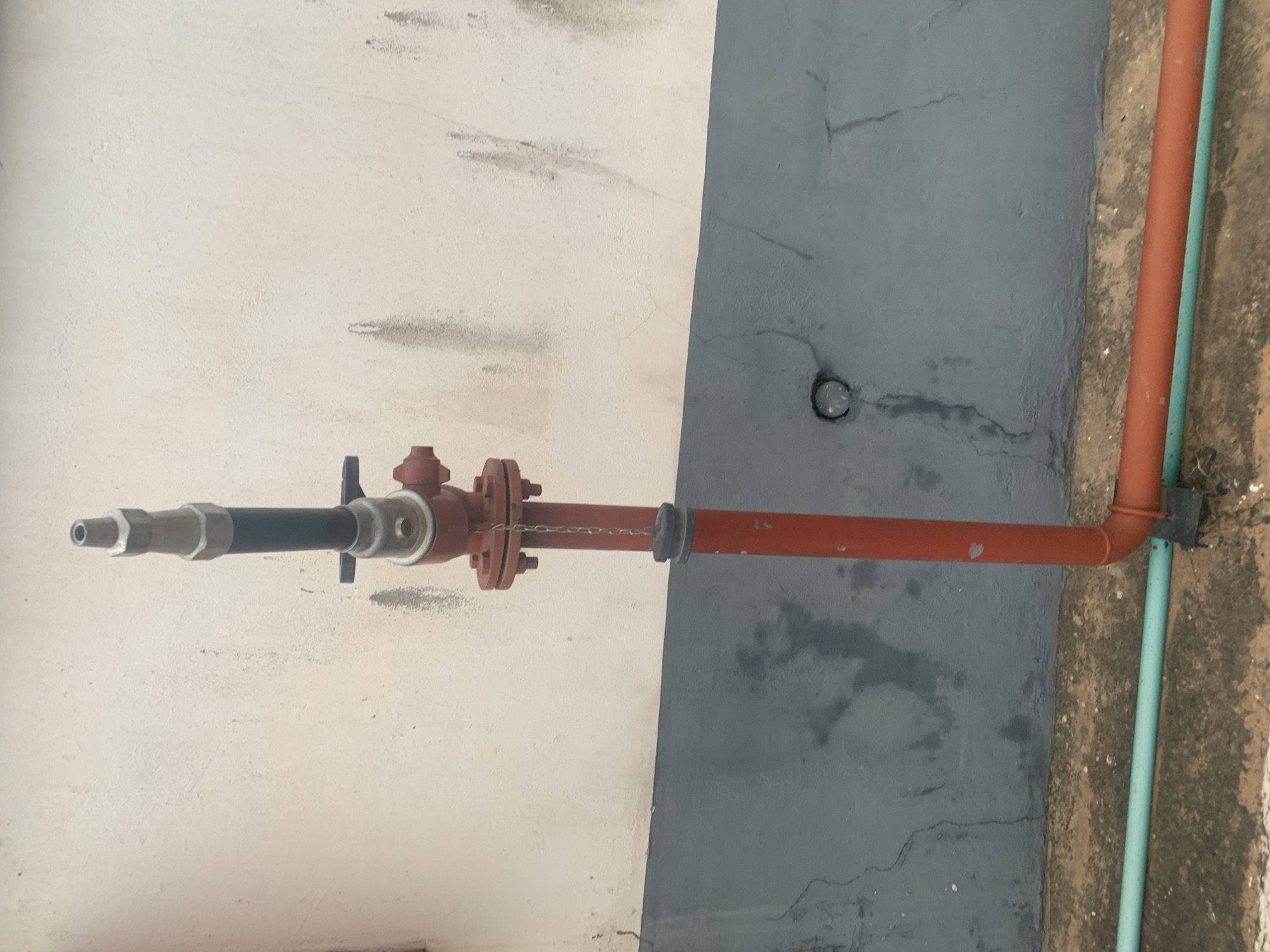 An outdoor, eye-level, medium shot captures a section of a building's exterior wall, divided horizontally by a gray band, with a reddish-orange pipe assembly protruding vertically from the ground. The upper portion of the wall is a textured, light-colored surface with faint streaks of dark discoloration. The lower portion of the wall is a darker gray and exhibits cracks. In the foreground, a light blue pipe runs horizontally at ground level, parallel to a reddish-orange pipe that bends at a right angle from a horizontal position to connect to the vertical pipe assembly. This assembly consists of several components: a lower red pipe section, a flanged connection with bolts, a red valve-like apparatus with a small red handle, a black cylindrical section, and a silver nozzle at the top. A thin metal chain connects the nozzle assembly to a black rubber cap attached to the lower red pipe section. The ground surface is visible at the bottom of the frame, appearing to be a mixture of concrete and dirt.