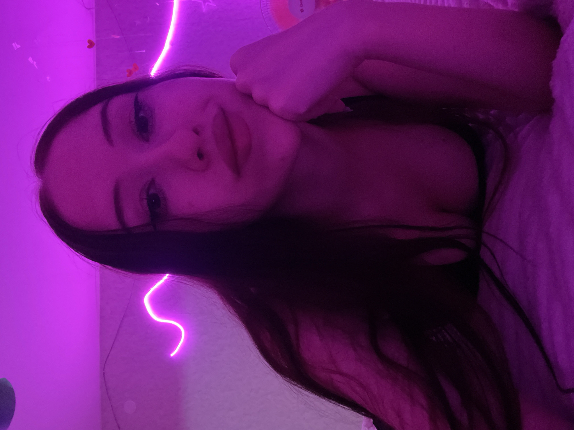 A young woman with long, dark hair is lying on a bed, illuminated by pink neon lights. She is resting her chin on her hand, looking towards the camera with full lips. Her chest is visible, and she appears to be wearing a dark top. The background is a textured wall with a wavy neon light strip and small heart decorations. There is also a blurred object that looks like a fan on the right side. The scene is indoors and appears to be a bedroom. There is no discernible information that can place this image specifically in Votkinsk, Russia.