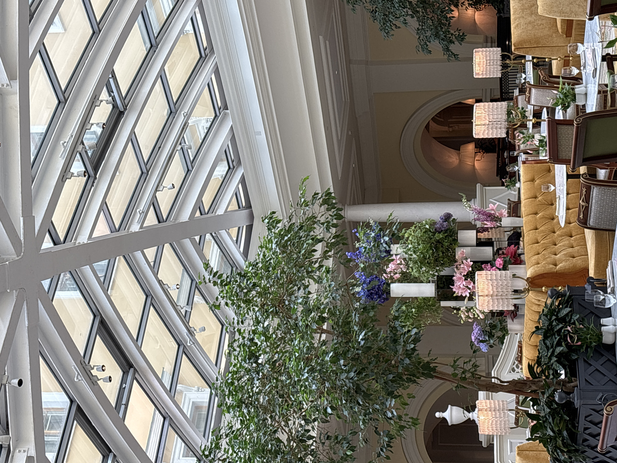 The image depicts an interior scene, likely a restaurant or event space, with a focus on elegant decor and architectural features. The most prominent elements are the large, diamond-shaped glass panes of a skylight, supported by a white metal framework. Sunlight filters through these panes, casting a warm glow on the scene below.
The setting is richly furnished with plush, mustard-yellow upholstered seating, appearing to be booths or banquettes. Tables are set with white tablecloths, glassware, and floral arrangements. The flowers are a mix of pink, purple, and white, adding a touch of color to the sophisticated ambiance. Two distinctive lamps with woven shades are visible, contributing to the warm and inviting atmosphere.
A large, leafy tree, likely artificial given its placement indoors and abundance of foliage, is positioned in the foreground, its branches reaching towards the skylight. Behind the tree and seating, architectural details like arched doorways and decorative molding are visible, suggesting a grand and historic building. The overall impression is one of refined luxury and a serene, possibly evening, setting due to the artificial lighting.
Given the location context of Saint Petersburg, Russia, one can infer this is a high-end establishment, possibly in a historic district. The image does not show any people, suggesting the activity might be a quiet moment before guests arrive or after they have left, or it's a view of a reserved area. No text is visible in the image.