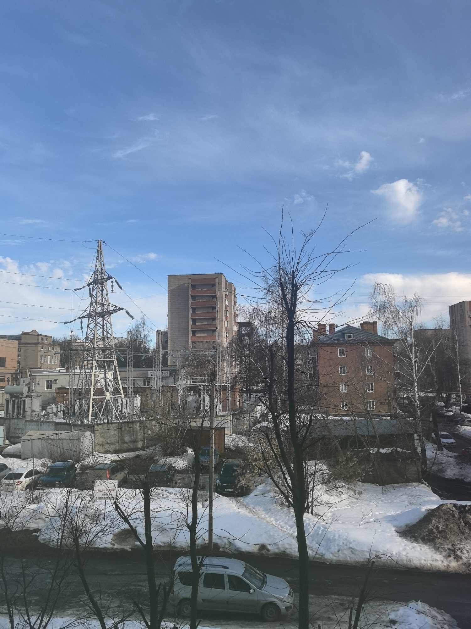 A wide-angle, elevated view captures an urban landscape in Sarapul, Russia, under a bright blue sky with sparse white clouds, indicative of a late winter or early spring day. The ground is largely covered in melting snow, revealing patches of dark earth and slush.
In the foreground, numerous bare deciduous trees, primarily with dark, slender branches, partially obstruct the view, with one prominent tree trunk rising centrally on the right. A dark asphalt road runs horizontally across the lower third of the frame. A silver compact SUV is parked on the left side of this road, partially visible.
The midground features a large electrical substation complex. A tall, lattice-steel transmission tower dominates the left-center, with numerous power lines extending from it and across the scene. Below the tower are grey industrial buildings, transformers, and insulators. To the right of the substation, a parking area contains several parked vehicles, including dark-colored SUVs and a white sedan. A partially visible white sign with Cyrillic text, reading "АГЕНТСТВО ЛОМБАРД Р" (Agency Lombard P) and "НЕДВИЖИМОСТЬ" (Real Estate), is present among the vehicles.
The background consists of various multi-story buildings. To the far left, a light-colored multi-story building with numerous windows is visible. Behind the substation, a tall, grey building with vertical reddish-brown facade elements stands prominently. To the right, a distinctive brick-red multi-story building with a dark grey gabled roof, two brick chimneys, and multiple white-framed windows is visible. Further to the right, another multi-story building is partially obscured by trees. No individuals are visible within the scene.