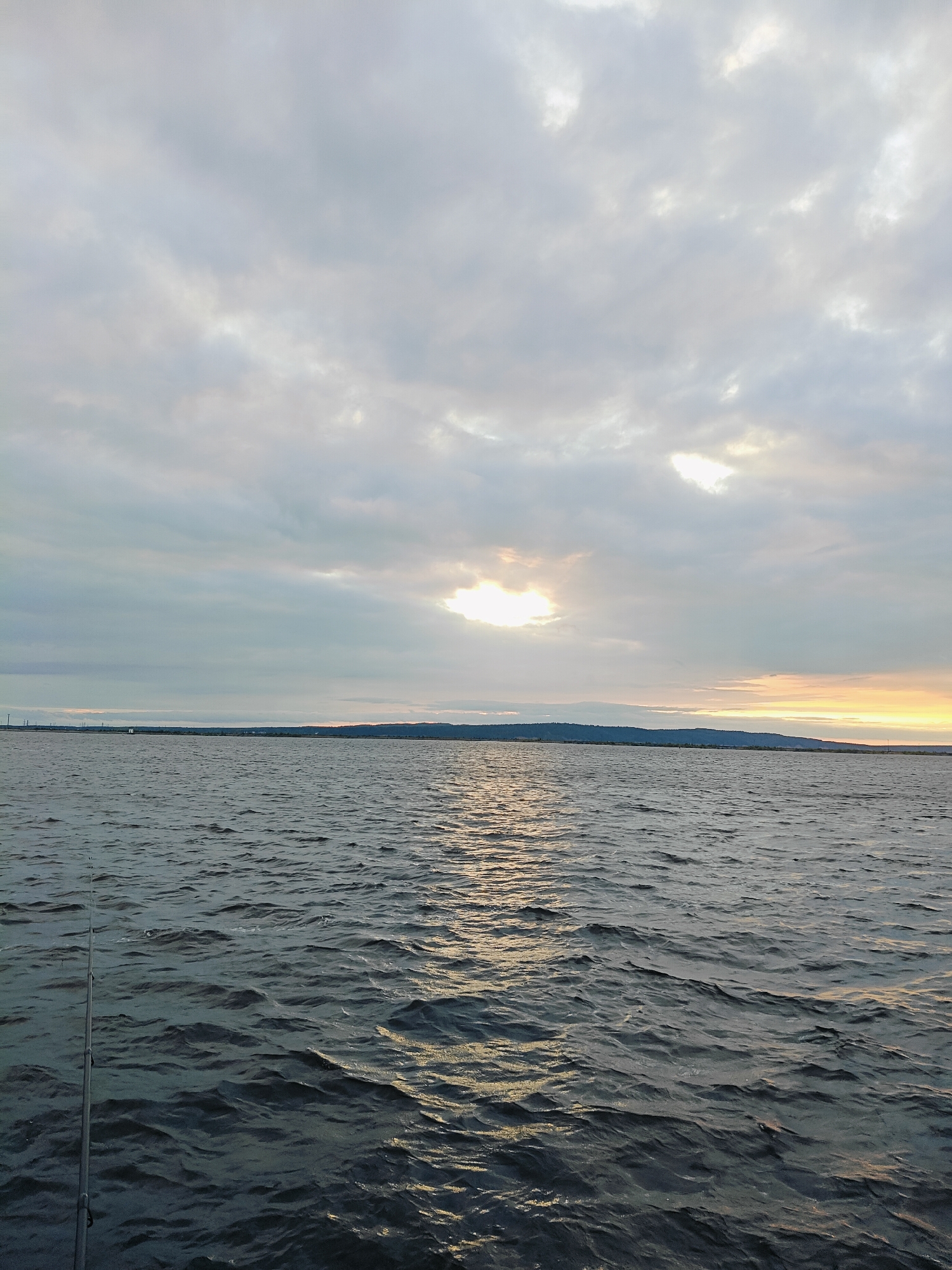 The image depicts a body of water, possibly a river or reservoir, under an overcast sky. A fishing rod is visible in the lower left corner, suggesting a fishing activity. The horizon line shows a distant shoreline with some vegetation or low hills. The sun is partially obscured by clouds, casting a diffused light and creating a shimmering reflection on the water's surface. The overall atmosphere is calm and serene. This scene could be located on the Kama River in Chaikovsky, Russia, a city known for its industrial presence and its location on the river.