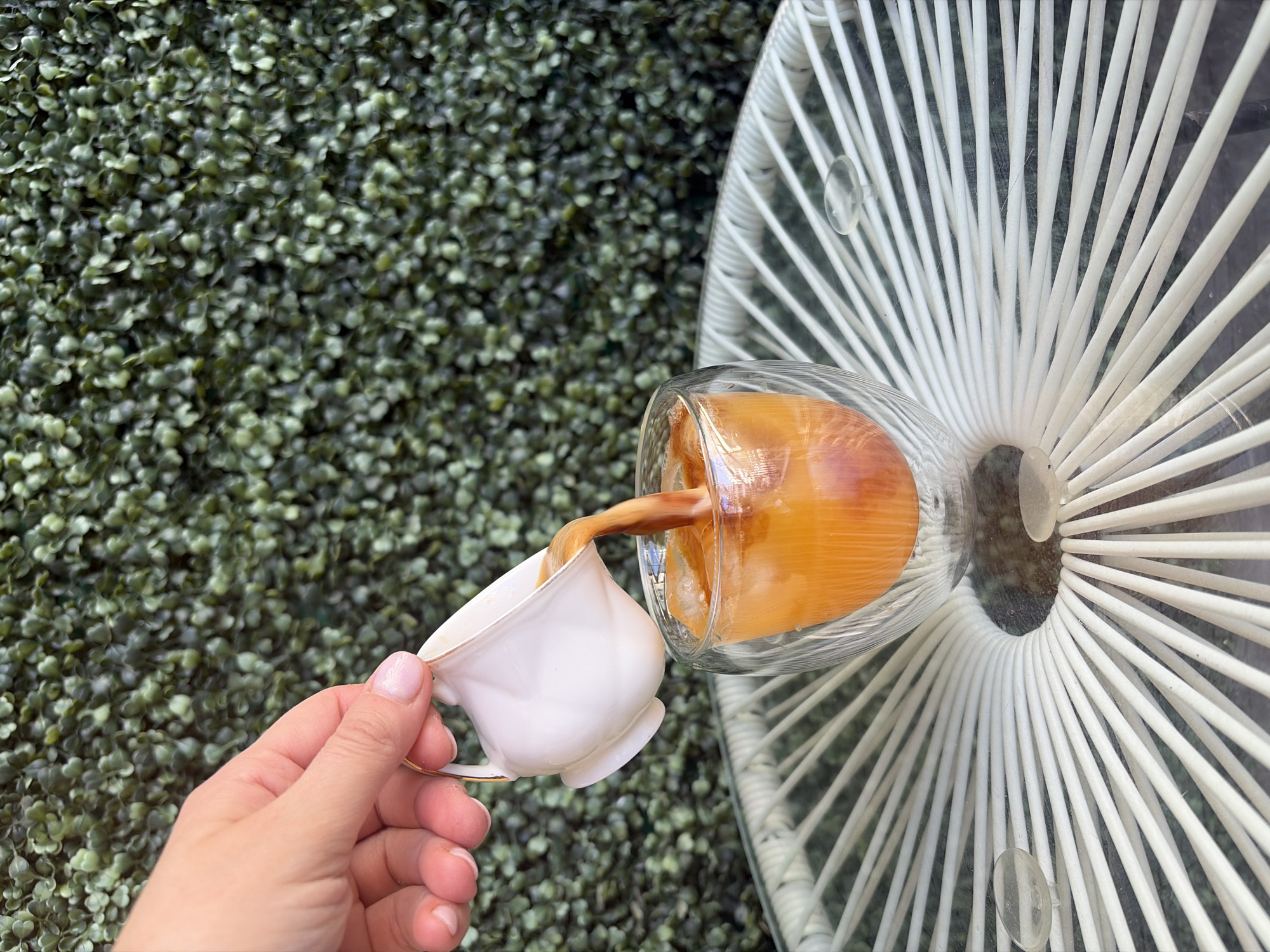 In Budva, Montenegro, a hand with manicured fingernails is seen pouring a dark liquid from a small white creamer with a gold rim into a clear glass filled with ice and an orange beverage. The glass is a double-walled design and sits on a white, circular table with a radial pattern of thin, woven strands. In the background, a dense green wall of artificial foliage serves as a backdrop. The scene captures a moment of beverage preparation, possibly coffee or a cocktail, against an outdoor or balcony setting.