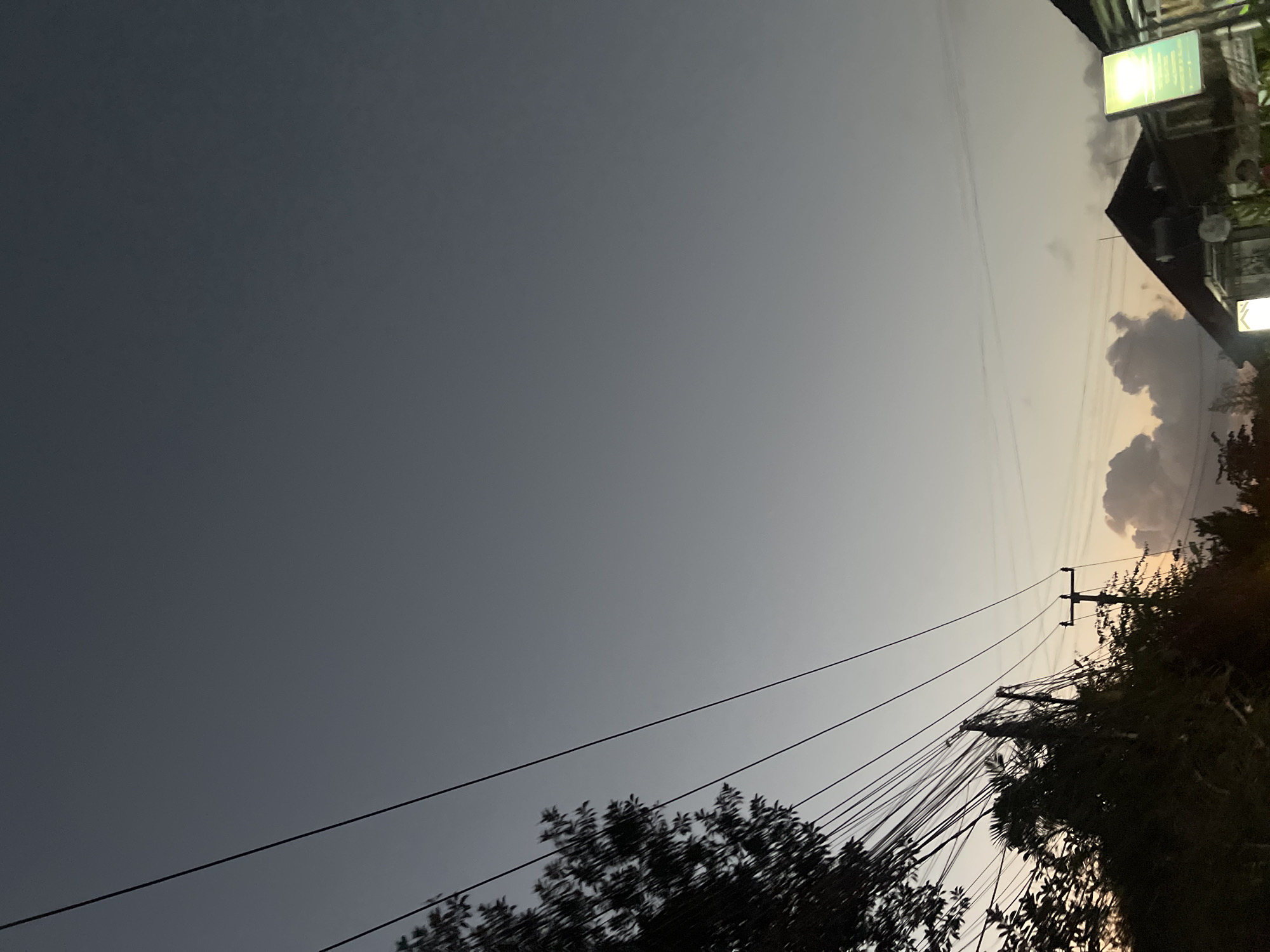 The image shows a view of the sky at dusk in North Kuta, Indonesia. Overhead, a network of dark power lines crisscrosses the frame, some strung between utility poles that are partially obscured by dense foliage and trees on the left side of the image. The sky transitions from a deep slate gray at the top to a lighter, pale gray and then a soft yellowish-orange hue near the horizon. A few scattered clouds are visible, particularly towards the right, illuminated by the fading sunlight. Silhouetted against the twilight sky are the dark shapes of building rooftops, and a brightly lit green sign is visible on the right edge of the frame, suggesting a commercial area. The overall impression is one of an urban or suburban street scene at the end of the day.