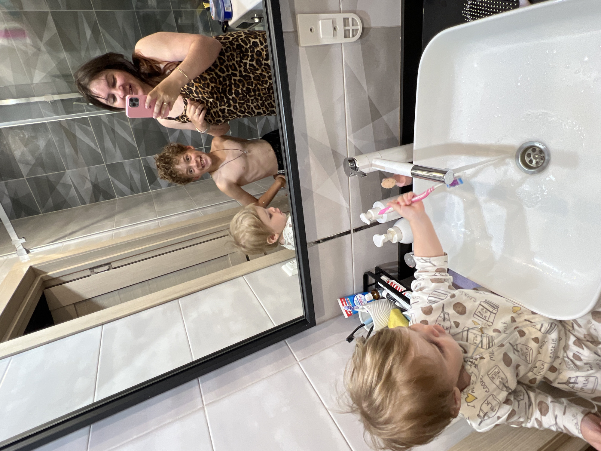 In a brightly lit bathroom, a woman wearing a leopard-print dress is reflected in a mirror, holding a pink smartphone and smiling. Beside her in the reflection, a young boy with curly hair grins at the camera. In the foreground, a toddler in a pajama onesie with a milk and cookie pattern is positioned at a sink. The toddler is holding a pink toothbrush, with water from the faucet running over it and into the sink. The bathroom features tiled walls, a modern sink and faucet, and various toiletries on the counter. The overall scene captures a candid moment of family life in a bathroom setting.