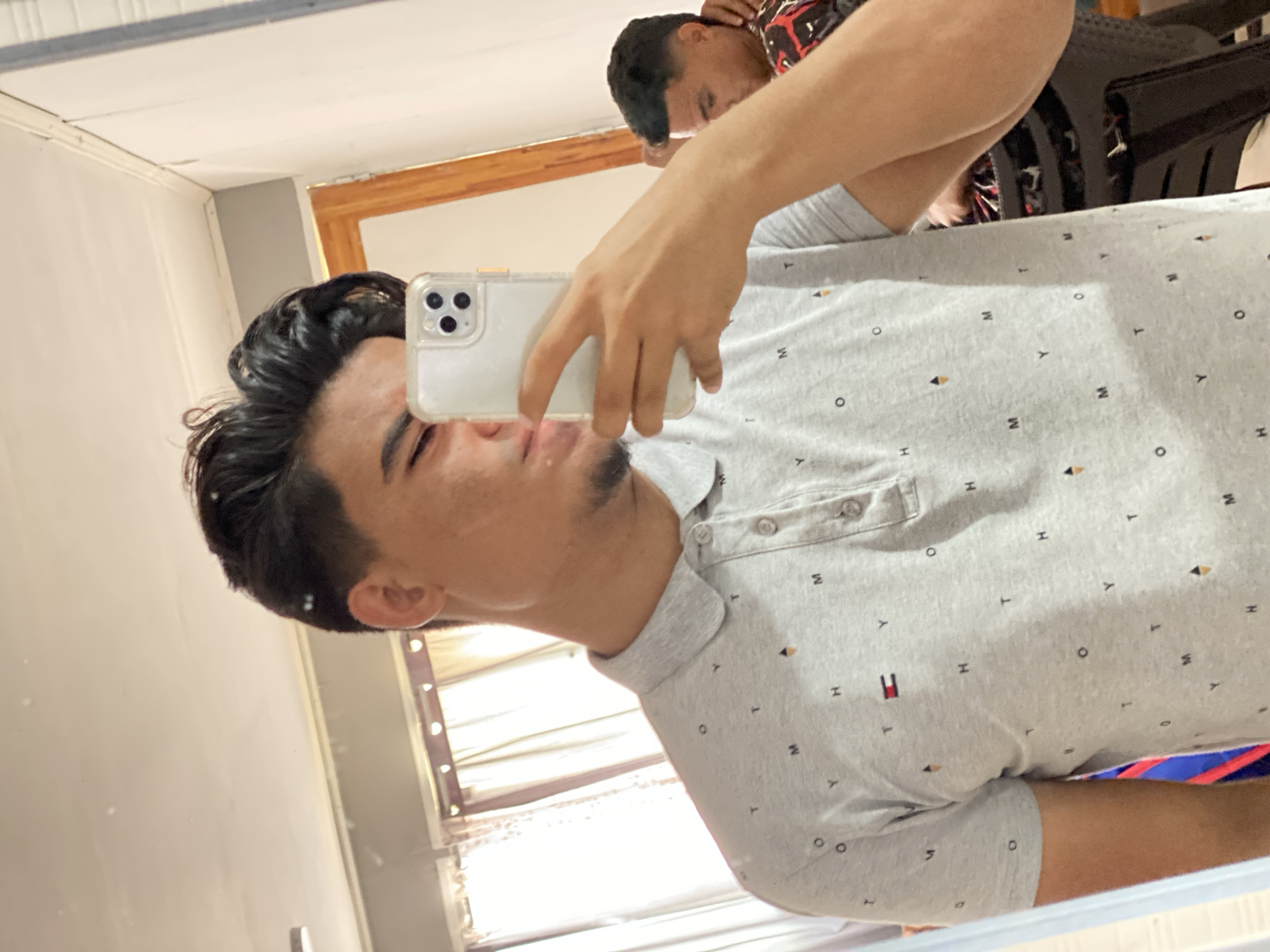 A young man is taking a selfie in a mirror. He is wearing a light gray polo shirt patterned with small "H", "Y", "M", "T", and "O" shapes, and a red and white Tommy Hilfiger logo. His dark hair is styled upwards. He is holding a white smartphone in his left hand. Reflected in the mirror is another person wearing a red and black patterned shirt. The scene is indoors, possibly a bedroom or living space. The exact location in Altagracia, Venezuela cannot be determined from the image alone.