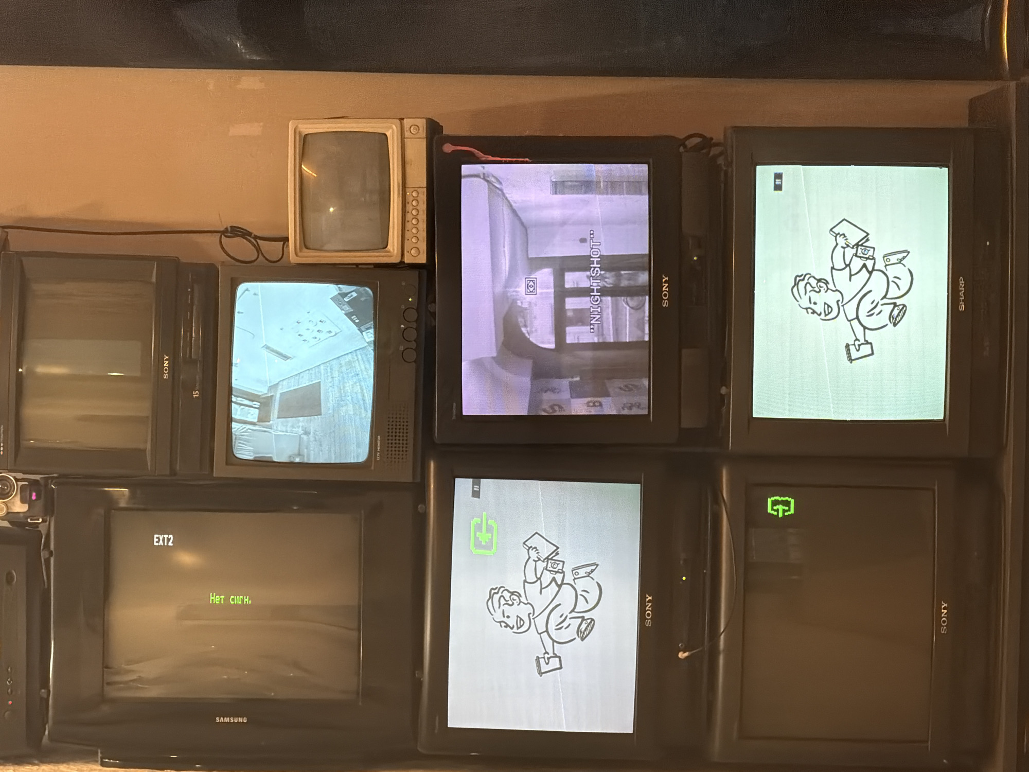 The image shows a collection of nine televisions stacked together against a wall. The TVs vary in size and brand, including models from Sony, Samsung, and Sharp. Some screens display video content, including what appears to be gameplay from a first-person shooter ("NIGHTSHOT" is visible), cartoon characters in a Fallout-style aesthetic, and a view of an interior space. One screen displays the text "EXT2" and the Russian phrase "Нет сигн.", meaning "No signal". The televisions are of the CRT variety, indicative of older technology. The environment is poorly lit and appears to be indoors. There is no information to suggest that the image was taken in Moscow, Russia.