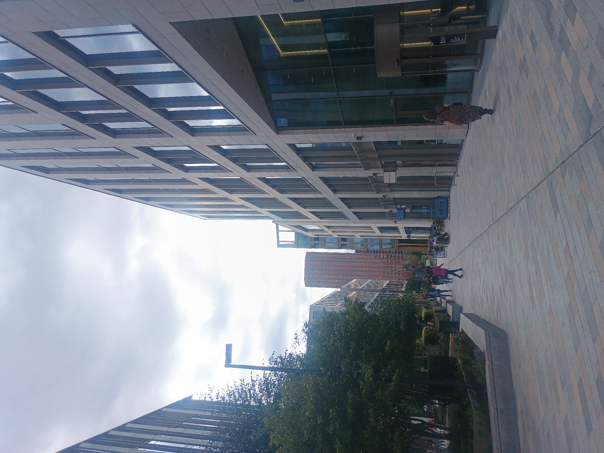In an urban setting, a modern architectural vista unfolds under an overcast sky. The scene is dominated by sleek, contemporary buildings with large windows reflecting the cloudy sky. On the right, a multi-story building with a light-colored stone facade features a glass entrance with a revolving door. A woman in a floral dress walks away from the camera towards the building entrance. On the left, another modern building with dark vertical accents lines the street. Between these structures, a pedestrian walkway paved with light-colored rectangular tiles stretches into the distance. Trees and landscaping with stone borders are visible on the left side of the walkway, creating a contrast between the built environment and nature. Several people are walking along the path, with a tall, reddish-brown cylindrical building visible in the background between the modern structures. A streetlamp with a unique angular design stands along the walkway. The overall atmosphere is one of a planned urban development with a focus on modern design.