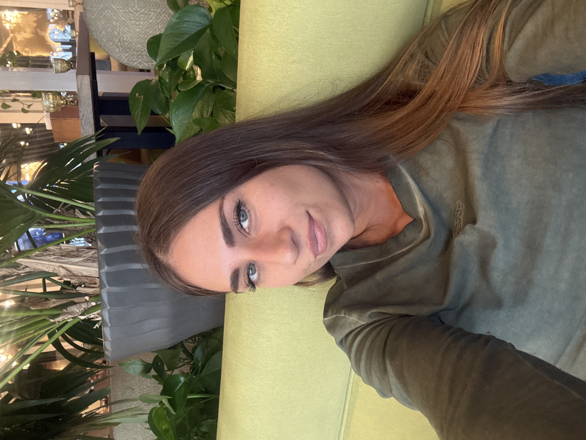 A medium shot captures a woman with long, brown hair and blue eyes looking directly at the camera. She is wearing a green long-sleeved shirt. The woman is seated on a lime green sofa. Behind her, a large gray planter holds a tall plant with long, thin leaves. Green vines spill out of the planter and hang down. In the background, a wooden table with a dark base holds two ornate gold goblets. The scene is indoors, with warm lighting suggesting it might be a restaurant or cafe. There is no specific indication that this location is in Komarovo, Russia.