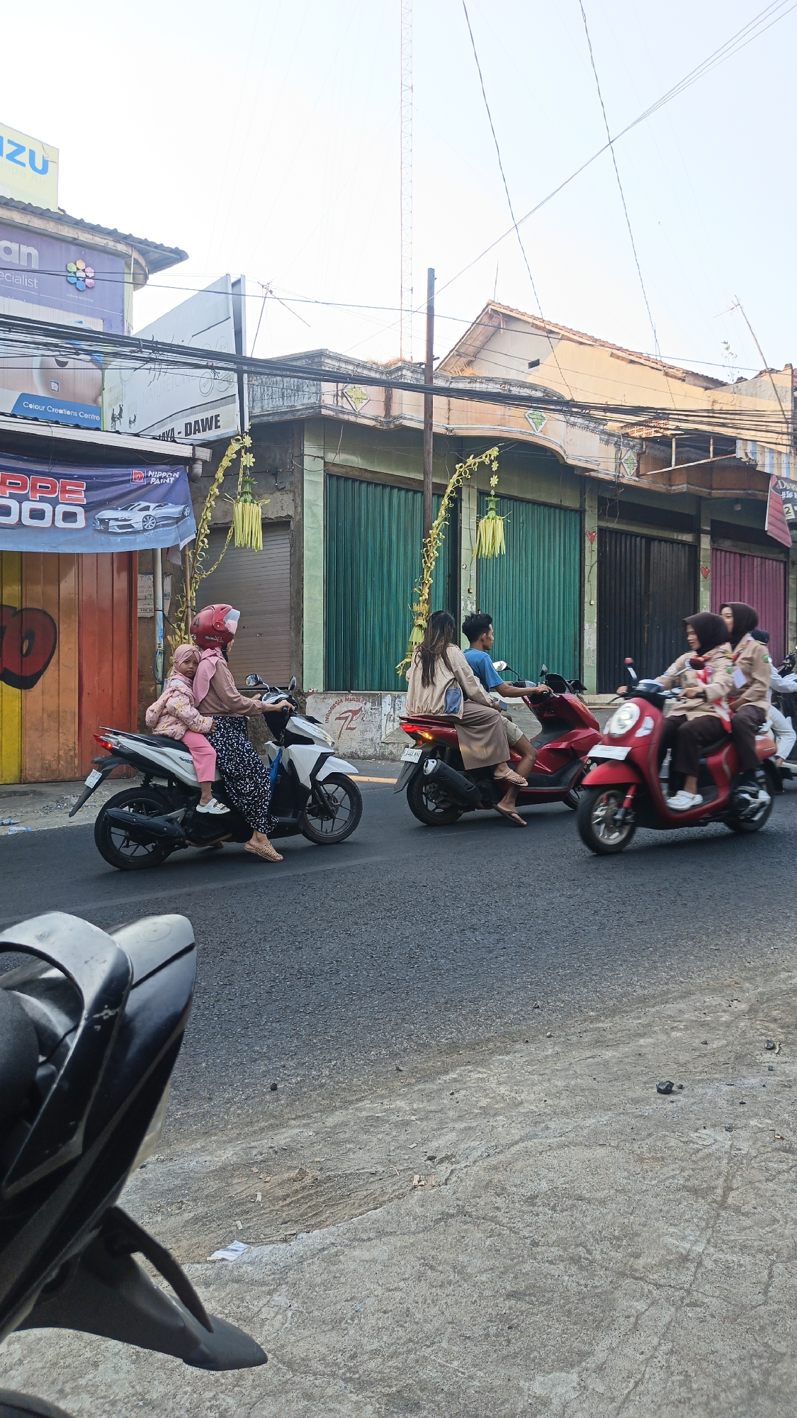 The image depicts a street scene in Dawe, Indonesia, featuring several individuals riding motorcycles. In the foreground, a motorcycle with a black rear section is partially visible. Moving further down the road, a woman in a pink hijab and a child in a pink jacket are riding a white scooter. The child is seated in front of the woman. To their right, a red scooter is carrying two people: a woman wearing a beige hijab and a man. Further to the right, another red scooter carries two young women wearing light brown hijabs and uniforms, possibly scout uniforms. The street is paved with asphalt, and there are buildings lining the street with green and red shutters. Overhead, power lines crisscross the sky, and a tall tower is visible in the background. Yellow decorative elements are hanging from poles along the street. The overall lighting suggests it is daytime.