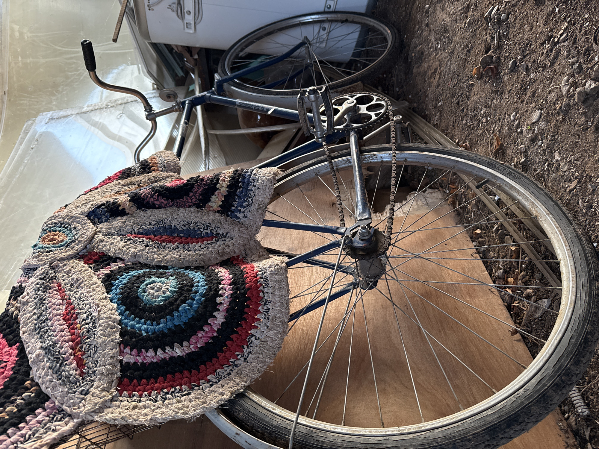The image shows a discarded blue bicycle partially covered with a colorful, circular rag rug. The bicycle is resting on a sheet of plywood near dirt and debris. The location is not discernible from the image.