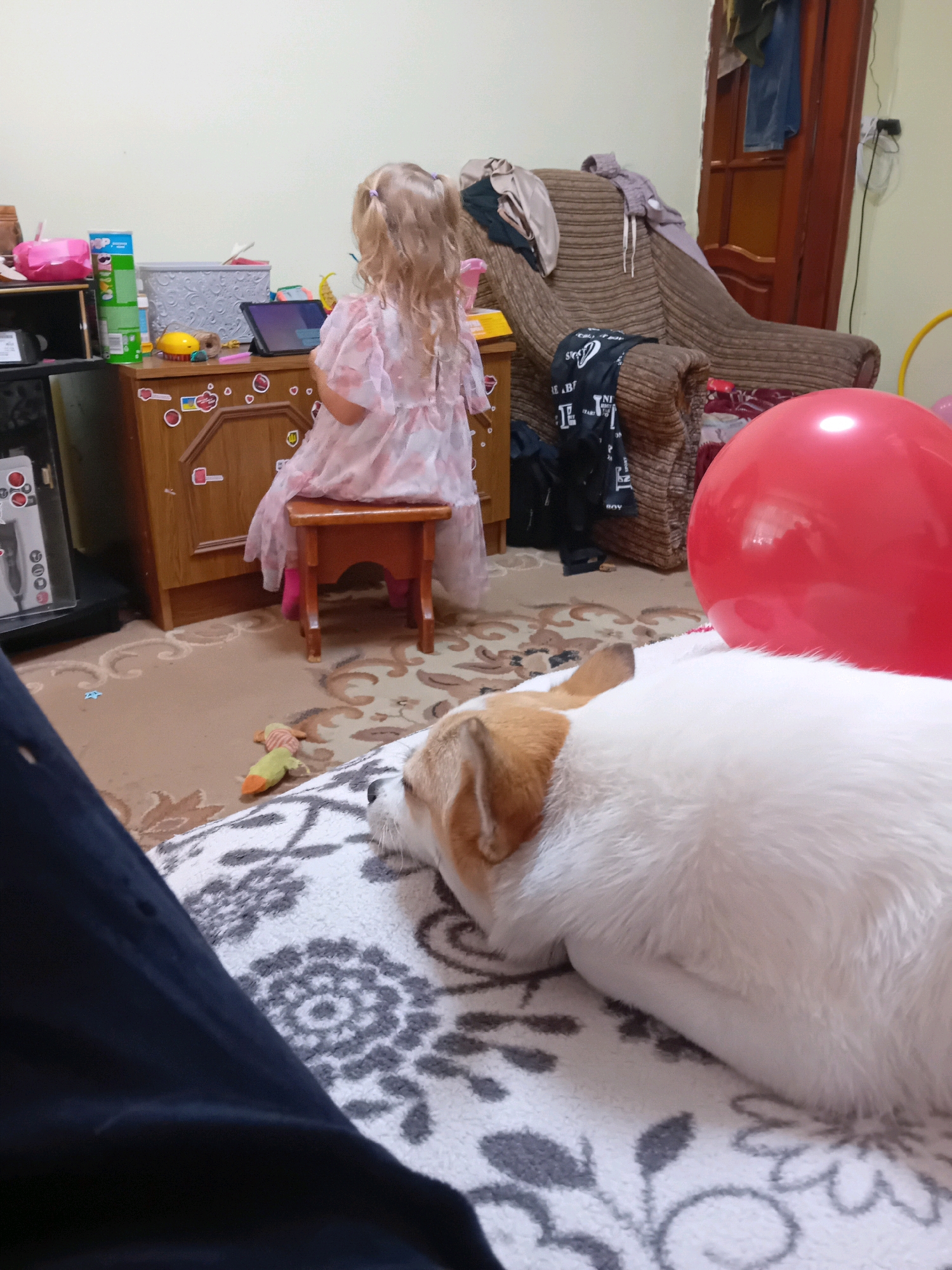 The image depicts an indoor scene, likely a residential living room, located in Chuhuiv, Ukraine. In the foreground, a white dog with tan markings is lying on a patterned rug. To the left and slightly behind the dog, a person's dark clothing is visible. In the mid-ground, a young girl with blonde hair in pigtails is seated on a wooden stool, facing away from the camera, and appears to be engaged with a tablet device placed on a dresser. The dresser is adorned with stickers, including a Ukrainian flag. Various items are scattered around the dresser, such as a tall green can, a pink container, and what appears to be a toy. In the background, a large red balloon is prominently displayed. Further back, a brown armchair with clothing draped over it is visible, and a wooden door with electrical outlets is present. The overall impression is a casual, lived-in domestic environment.