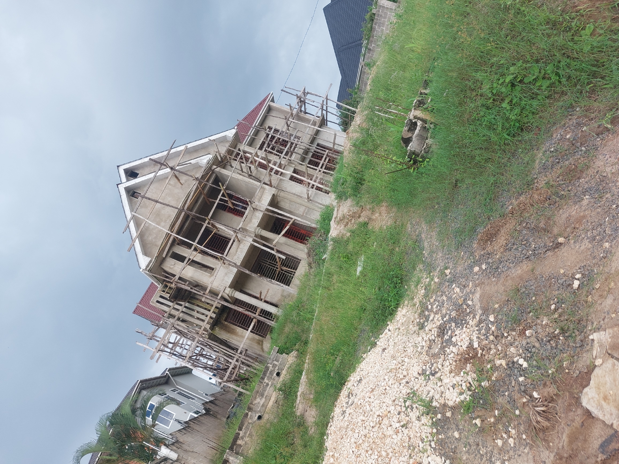 The image, which appears to be rotated 90 degrees clockwise, depicts a construction site within a residential area, likely in Dar es-Salaam, Tanzania. The central focus is a multi-story building, presumably a house or small apartment block, currently under construction. Its concrete structure is prominently surrounded by extensive wooden scaffolding. The building features a distinctive red tiled roof and multiple window openings, which are currently fitted with simple metal grates or bars.
To the lower left, another completed or nearly finished modern house is visible, distinguished by its grey exterior walls, white-framed windows, and a dark roof. A vibrant green palm tree stands near this residence. In the upper right background, part of another building with a dark-colored roof can be seen.
The foreground consists of uneven ground, presenting a mix of lush green grass, patches of light-colored gravel, and exposed dirt. Some concrete debris or blocks are scattered on the right side of the foreground. The sky above is overcast and grey, indicating cloudy weather with no direct sunlight. No people are visible, and there is no discernible text within the image.