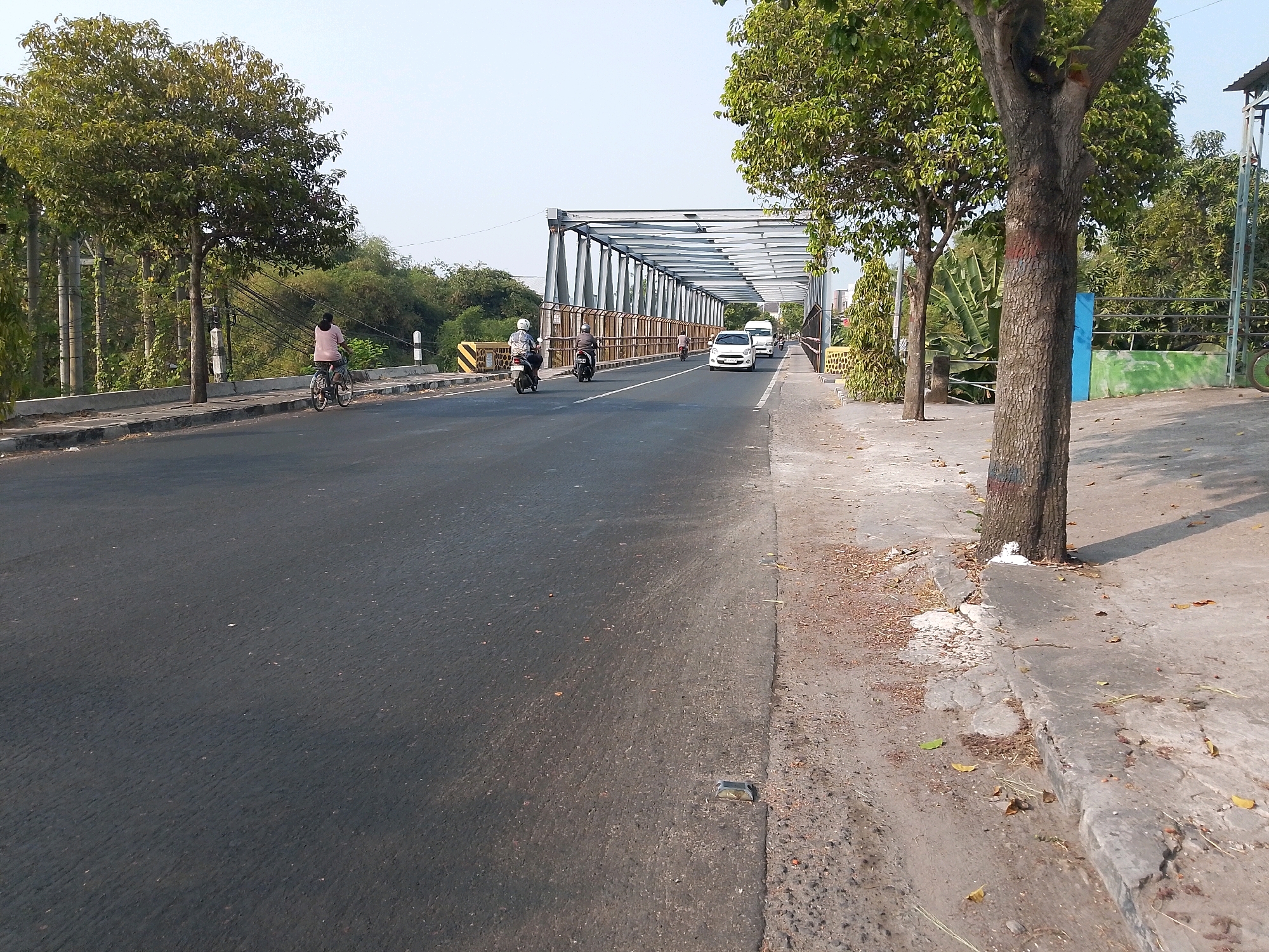 The image depicts a scene on a two-lane road leading onto a metal truss bridge, likely in Ngawi, Indonesia. The road is paved with asphalt and has white lane markings. On the left side of the road, a person is riding a bicycle, wearing a pink shirt. Further ahead, on the bridge, two motorcycles are in motion, with riders wearing helmets. There are also two white cars traveling on the bridge.
The bridge is supported by a robust metal structure. On the sides of the road and bridge, there are sidewalks made of concrete. Several trees with green foliage are present, one large tree is prominently featured on the right side of the image, with a textured bark and some red and blue markings near its base. The environment appears to be somewhat rural or suburban, with greenery and trees lining the road and surrounding areas. The sky is clear and bright, suggesting daylight. A small blue and green fence is visible to the right of the large tree.