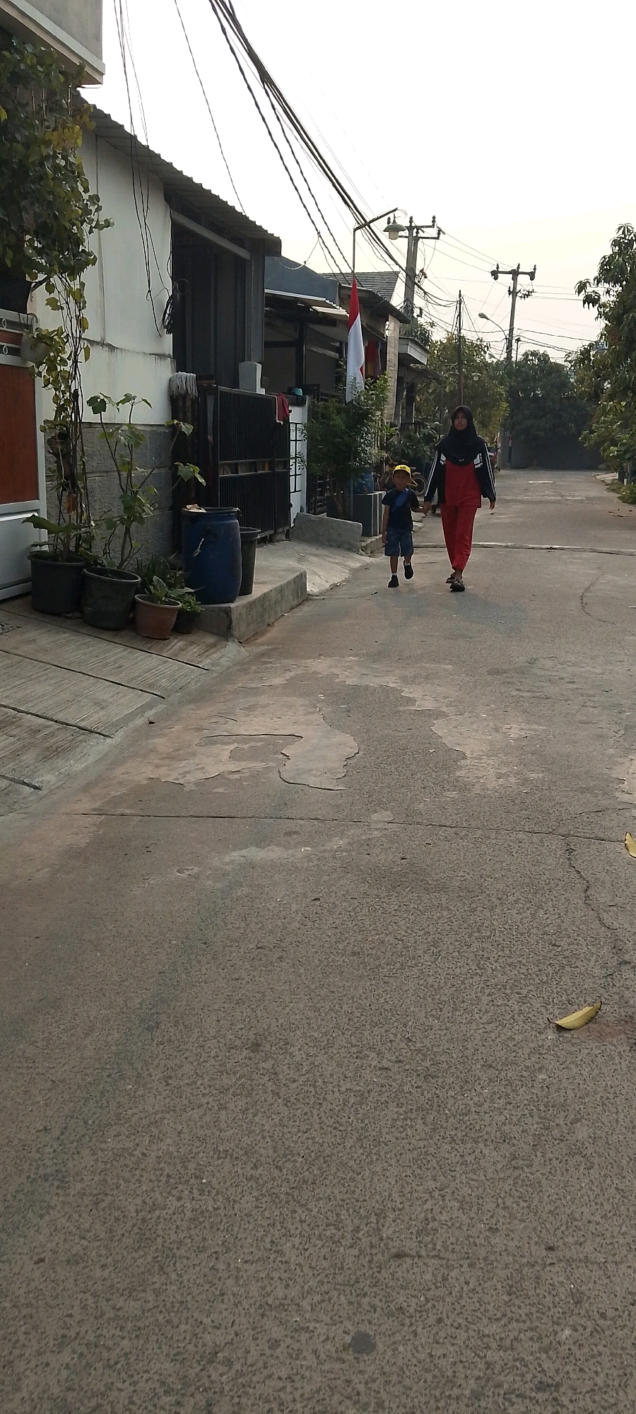 The image depicts a street scene in Satriamekar, Indonesia. On the right side of the frame, a woman wearing a black hijab and a red outfit walks hand-in-hand with a young boy in a blue shirt, blue shorts, and a yellow cap. The boy is on the left, slightly ahead of the woman. They are walking away from the viewer down a paved residential street. On the left side of the street, a two-story building with white walls and a corrugated metal roof is visible, with plants in pots along its base. The environment appears to be daytime, with overhead power lines and utility poles visible in the background. An Indonesian flag is displayed on a pole in front of one of the buildings.