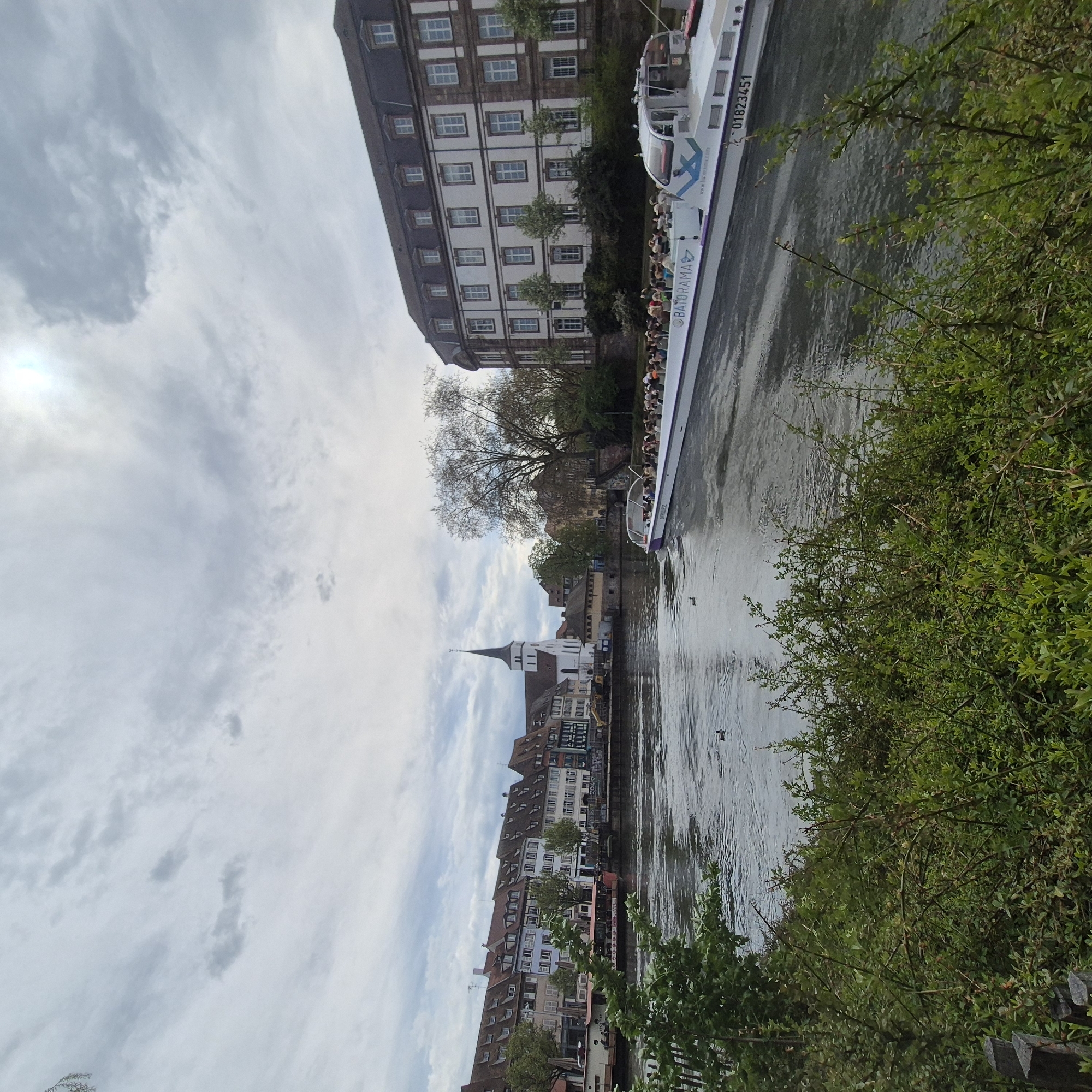The image depicts a river scene in Strasbourg, France, likely the Ill River. A tour boat, labeled "BATOBRAMA" and displaying the phone number "01823451", is navigating the river, filled with tourists. The river is flanked by buildings, mostly residential with some commercial establishments, characterized by traditional European architecture with pitched roofs and multiple stories. A church steeple is visible amidst the buildings. Vegetation, including trees and bushes, lines the riverbank. The sky is overcast with a mix of clouds.
