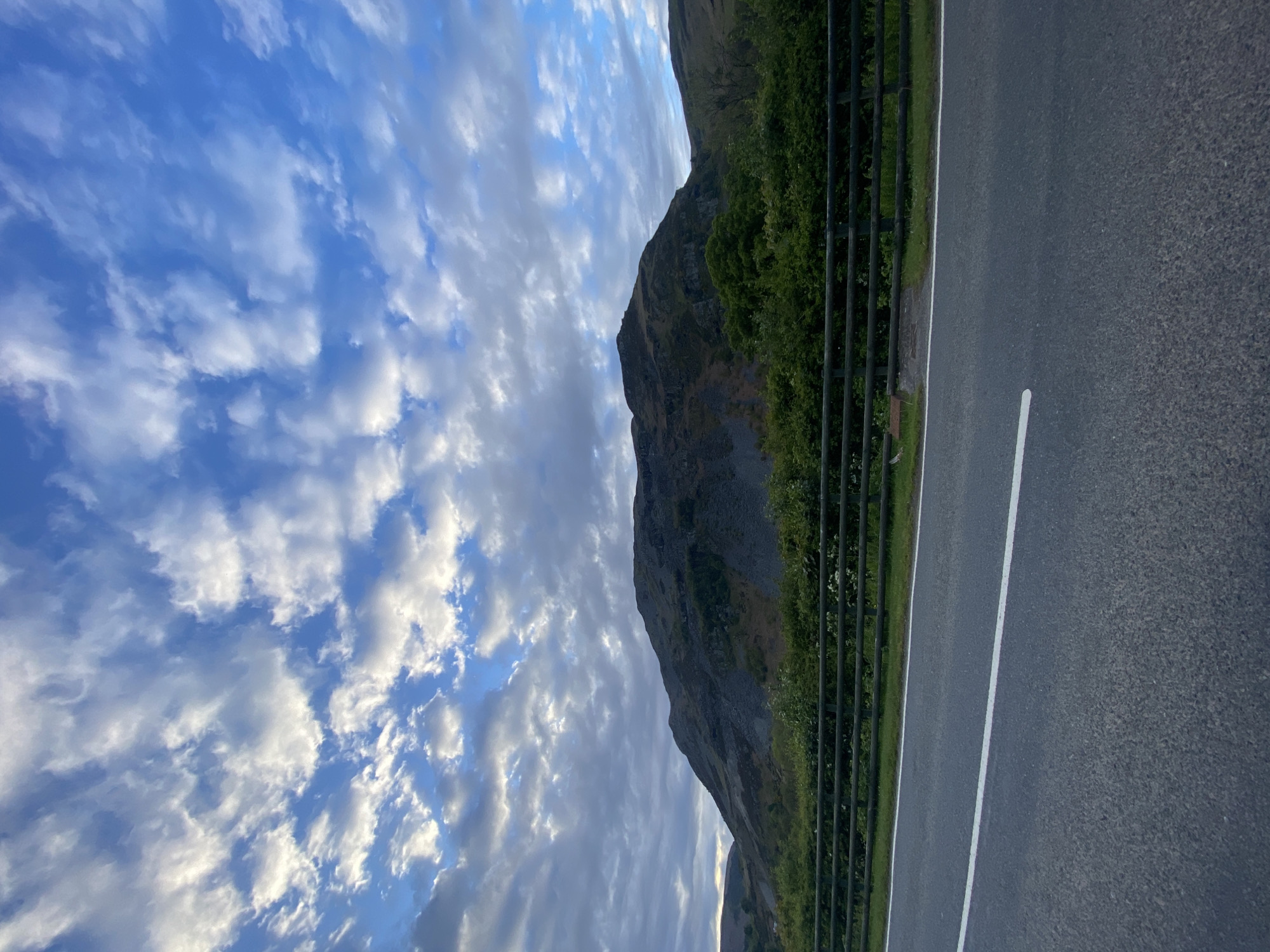 The image captures a scene in Llangynog, United Kingdom. The frame is divided into three primary sections. On the left, a cloudy blue sky fills the majority of the space. The sky is dynamic with varying shades of blue and white. Adjacent to the sky is a mountain partially covered in greenery, sloping downwards. A fence runs along the mountain. To the right of the fence is a paved road with a white lane marker. The road occupies the remaining portion of the image. The perspective is slightly angled, giving a view across the landscape. No people are visible.