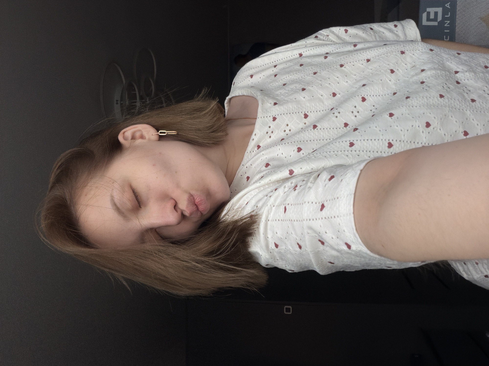 A young person, likely female, is depicted lying down indoors in Reshetnikovo, Russia. The individual's head is turned to the left, and their eyes are closed, with lips playfully pursed in a "kissy face" expression, suggesting a relaxed and casual moment.
They have light brown, shoulder-length hair and are wearing a white short-sleeved top. The top features a pattern of small red hearts and intricate embroidered eyelets, giving it a delicate, textured appearance. A thin, gold-colored chain necklace is visible around their neck, and a unique gold-toned earring, shaped like two interconnected oval links, is worn in their right ear.
The background is primarily dark, suggesting a dimly lit room or dark-colored furnishings. In the upper right portion of the image (when oriented as seen), part of a gray object is visible, displaying white capital letters and a symbol. The visible text includes an "E," a symbol resembling a stylized "P" or open square, and then "CINLA," possibly part of a brand name or logo. Faintly in the upper left, some curved metallic structures, possibly from a light fixture, can be seen against the dark wall. The scene captures an intimate and tranquil atmosphere.